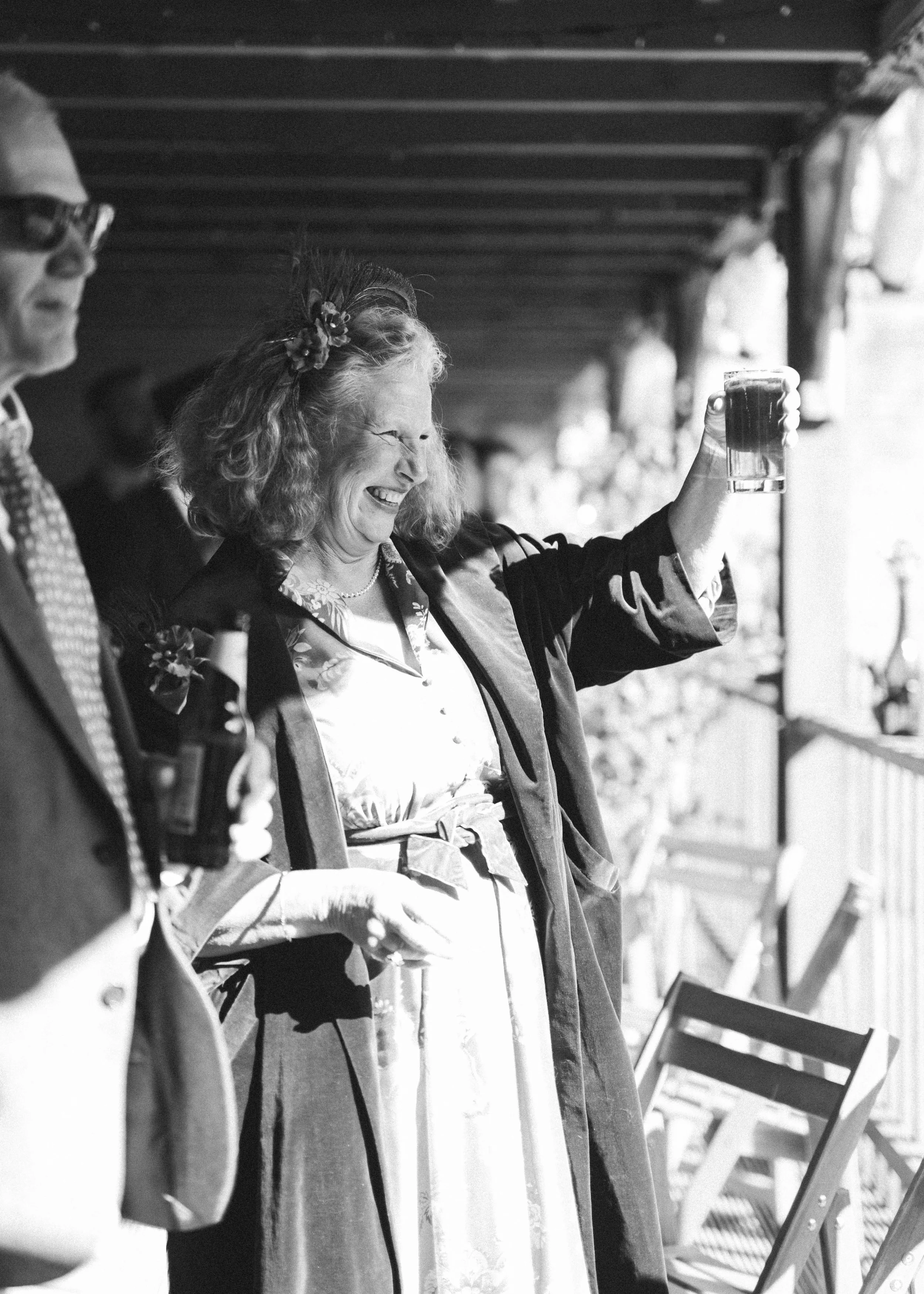 A woman with curly hair and a floral headband holding a glass of beer and smiling at an outdoor gathering.