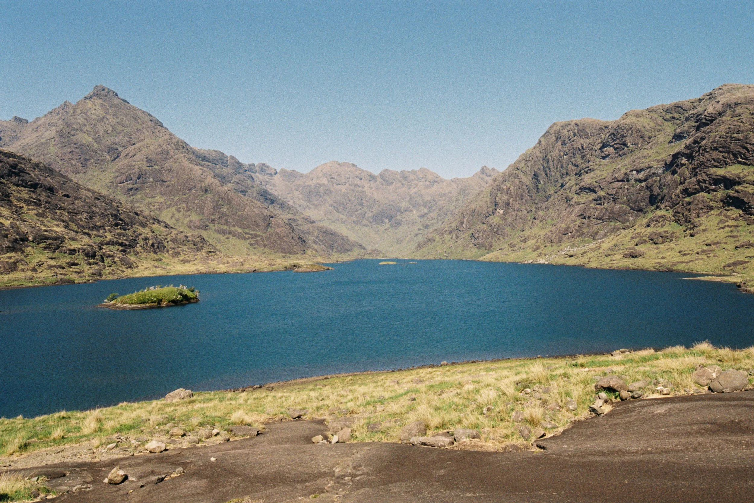 Scenic view of a large body of blue water surrounded by green grassy areas and rugged mountains under a clear blue sky.