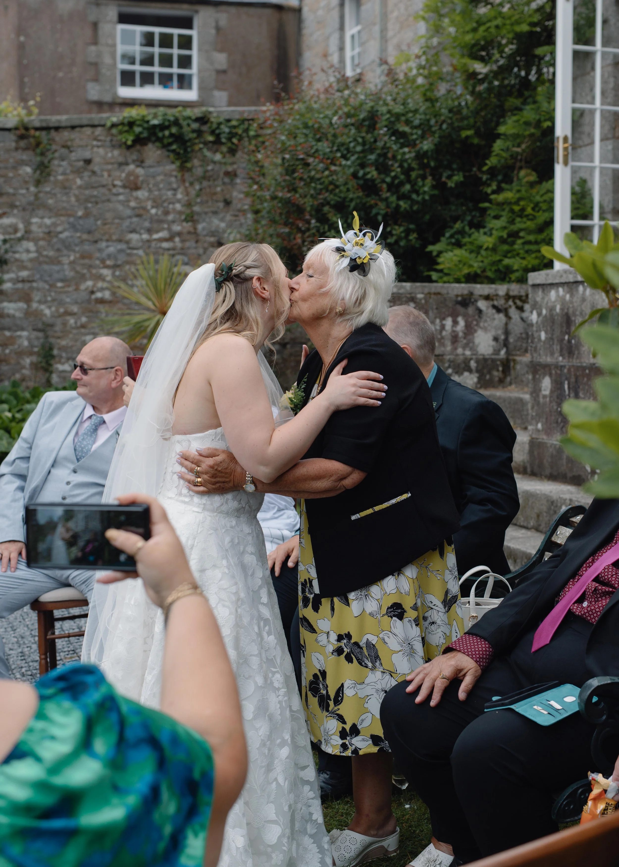 A wedding ceremony in a garden with a bride and an older woman sharing a kiss. The bride is in a white strapless wedding dress with a veil, and the older woman has white hair with a floral headpiece and is wearing a black jacket and a yellow floral d