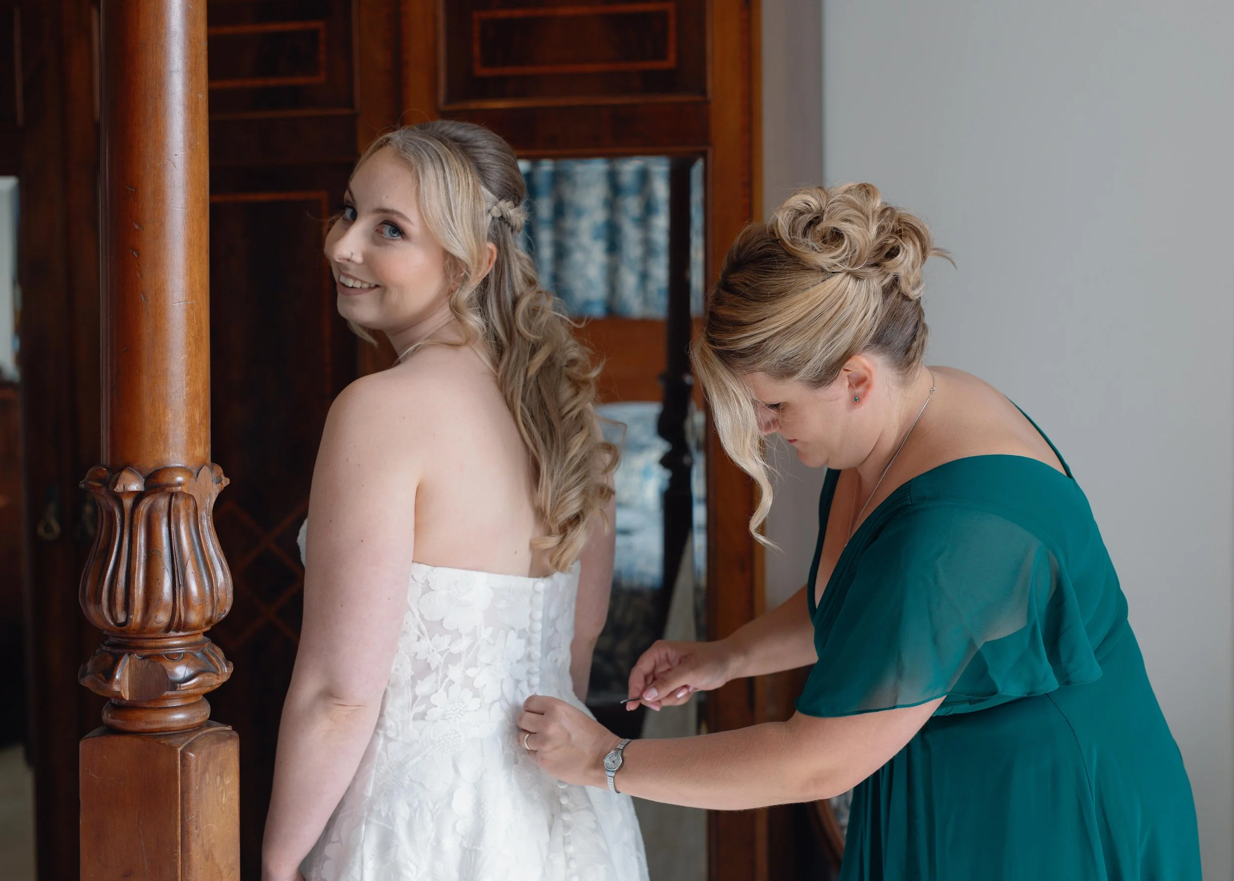 A bride in a white lace wedding dress is smiling while a woman in a green dress helps her with her dress, in a room with wooden furniture.