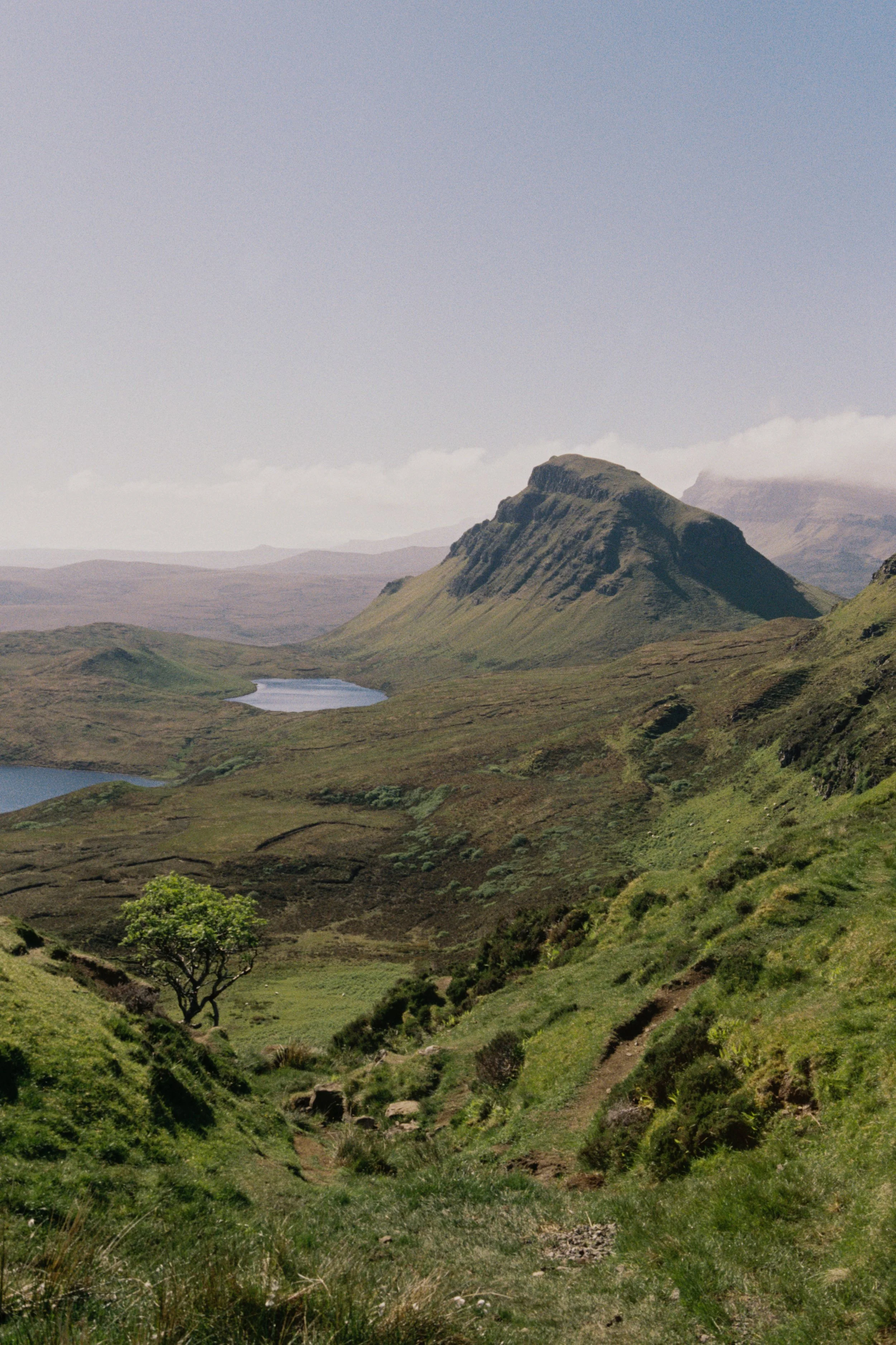 A scenic view of a mountainous landscape with a prominent mountain in the background, green rolling hills, a small lake, and a clear sky.