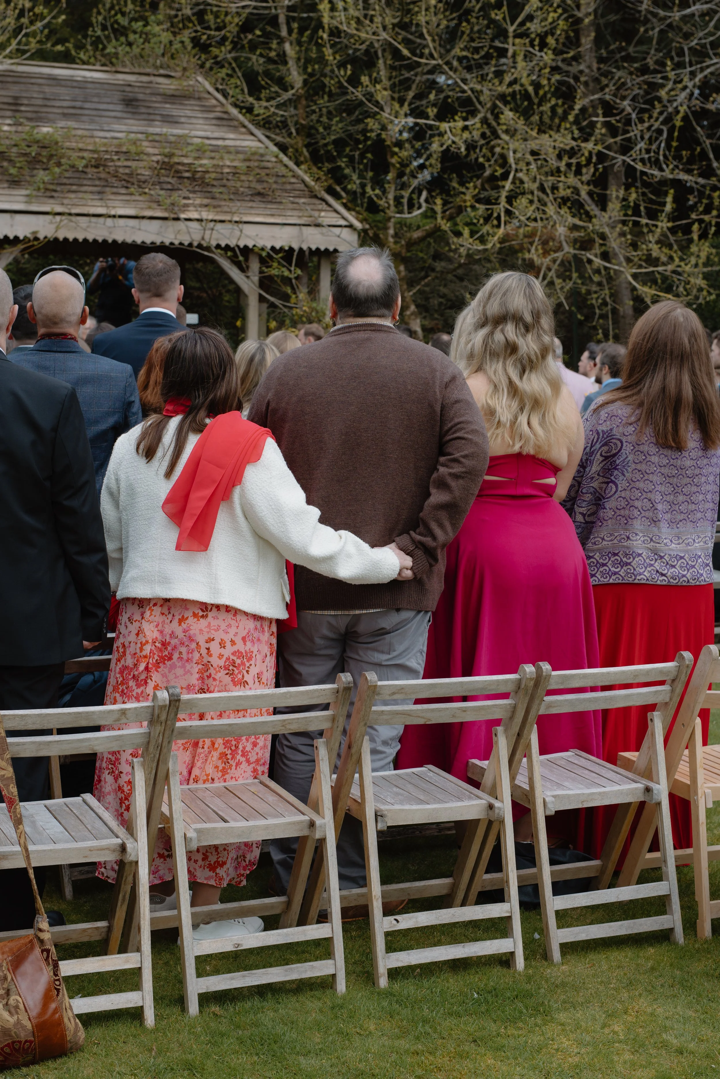 A group of people standing outdoors at a gathering, with some holding hands or embracing, under a tree and near a rustic wooden structure.