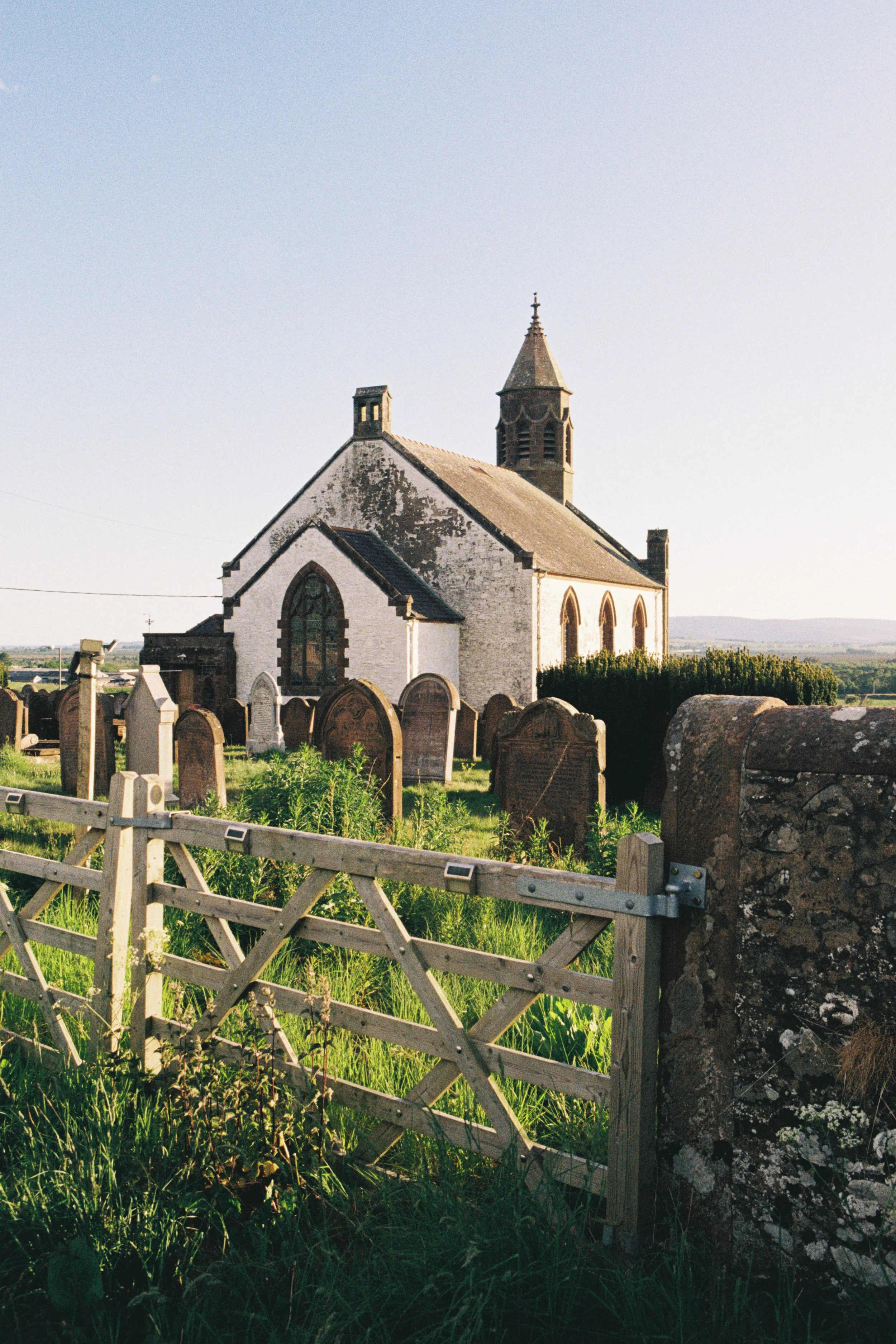 A small white church with a steeple and stained glass window, surrounded by a graveyard, green grass, and a wooden gate, with a clear sky and distant landscape in the background.