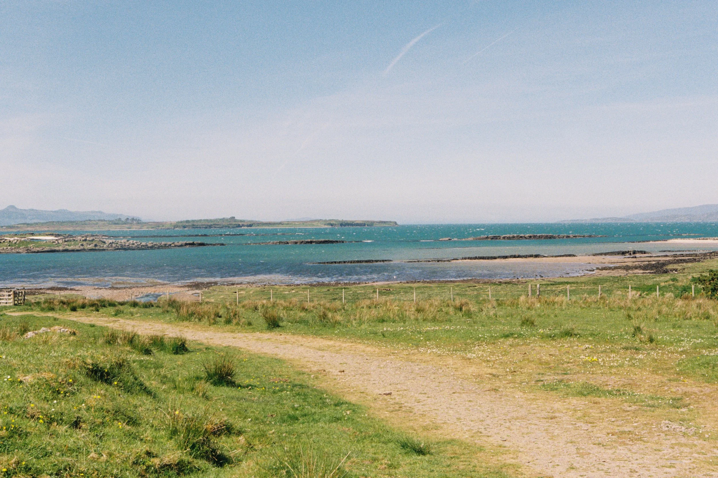 A scenic view of a coastal area with grassy paths, sandy shores, and turquoise ocean water, with distant hills and a clear blue sky.