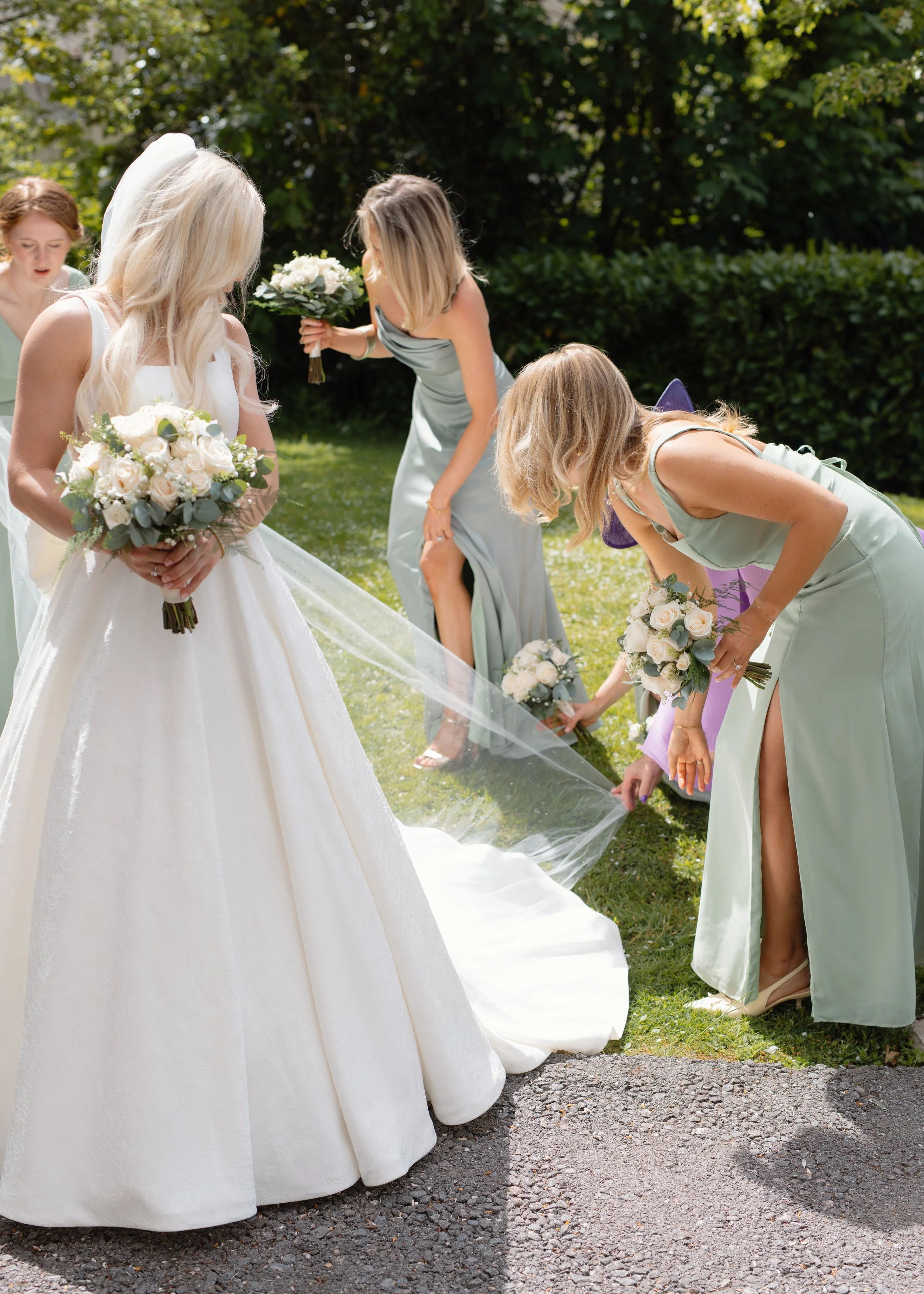 Women in formal dresses, including a bride in a white gown, adjusting the bride's train with bouquets of white roses and greenery outdoors on a sunny day.