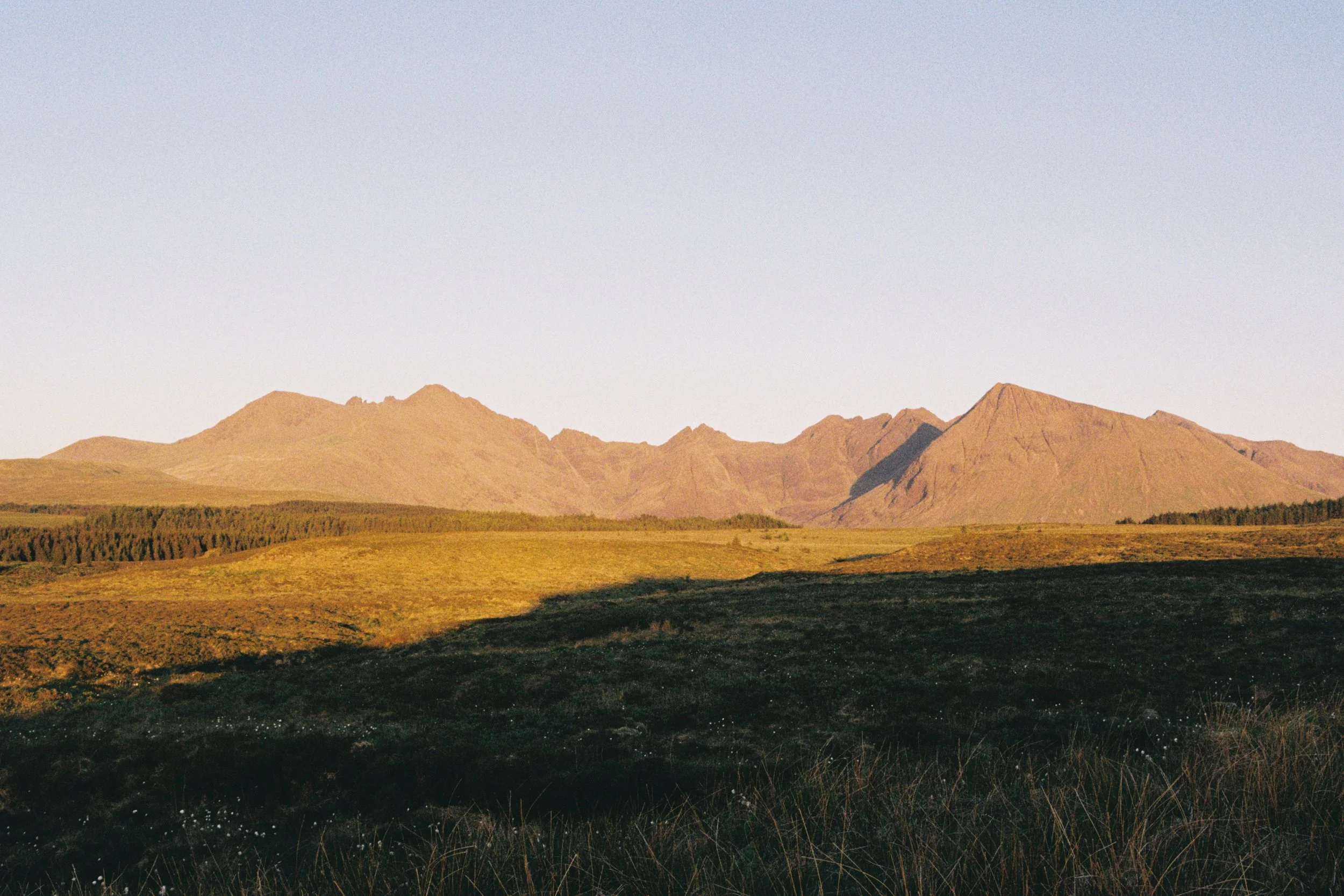 Scenic landscape with rolling hills and mountains under a clear sky, with shadows cast on the terrain, during late afternoon or early evening.