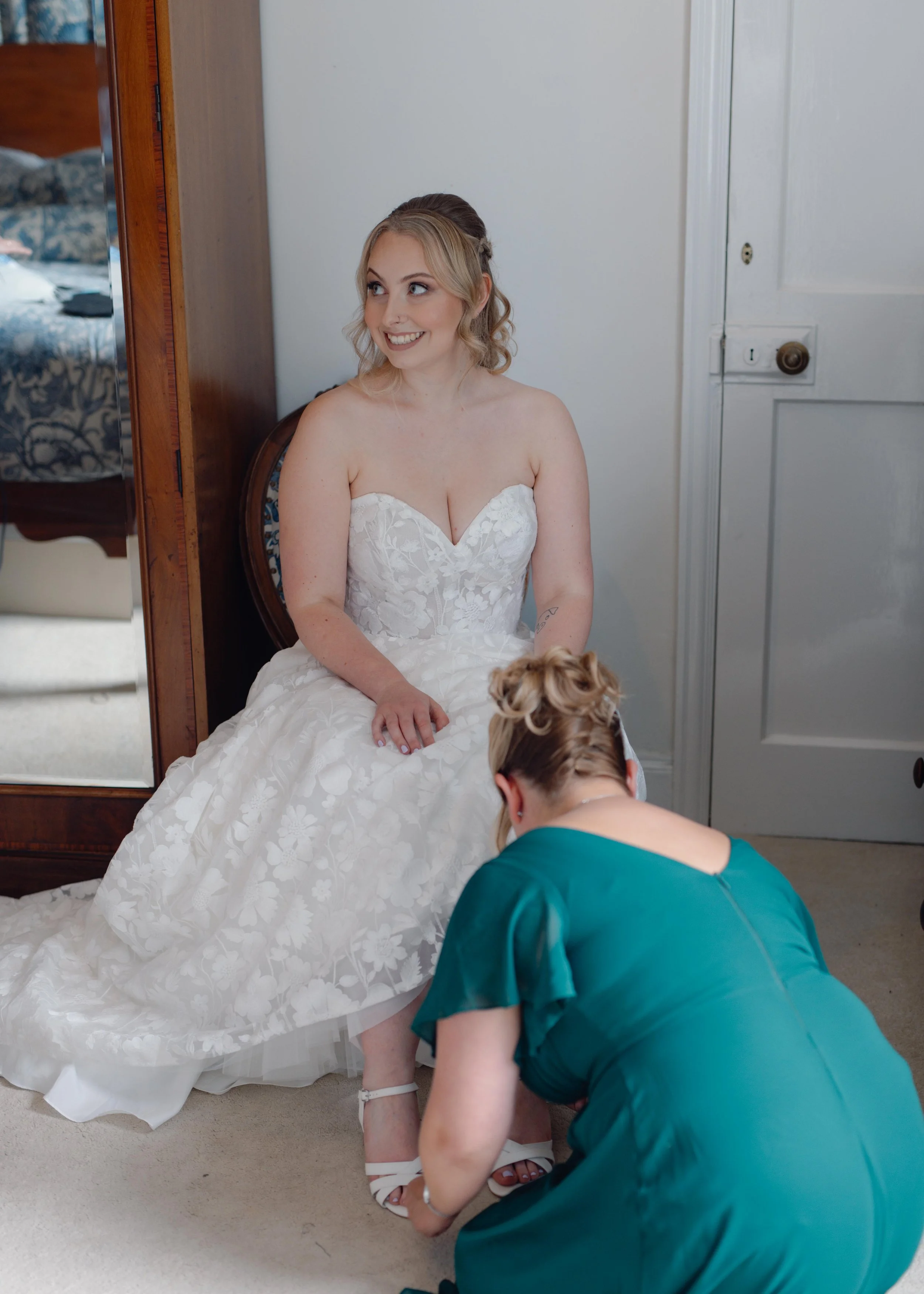 A bride in a white strapless wedding dress sits on a chair while a woman in a teal dress helps her put on white high-heeled shoes.