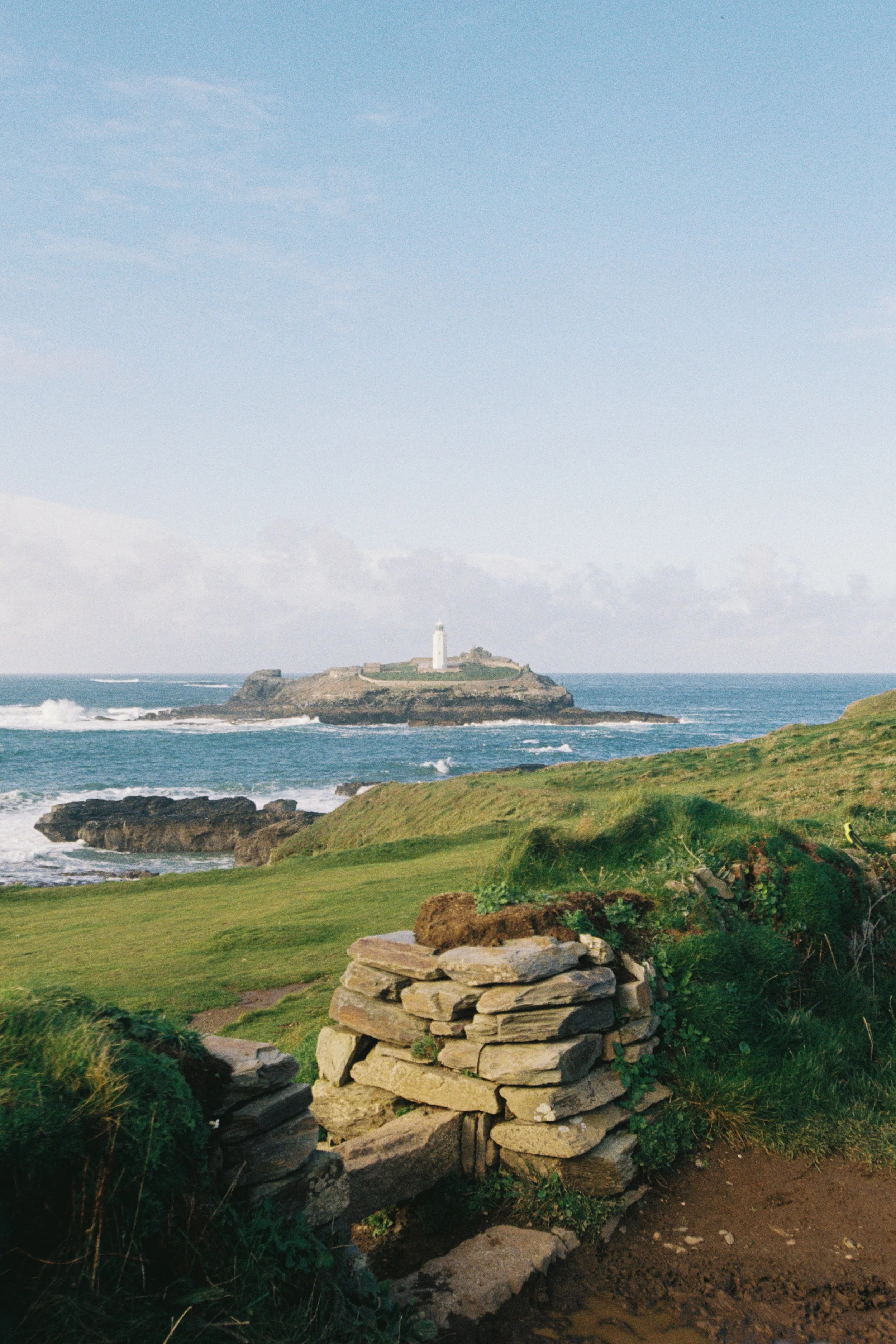A coastal scene featuring a stone wall in the foreground, green grassy areas, and a rocky shoreline with waves. A lighthouse is situated on a small island in the background under a partly cloudy blue sky.
