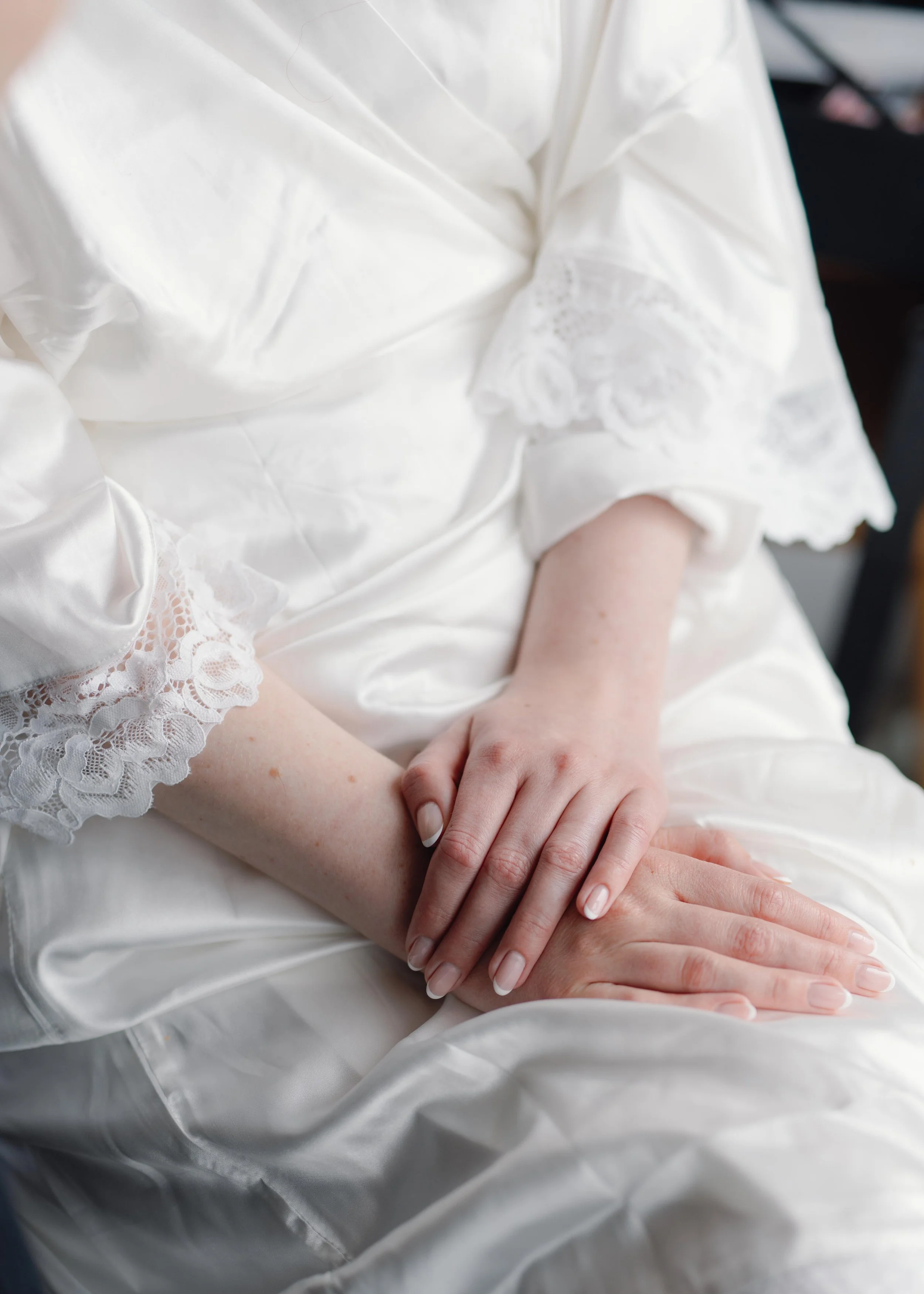 Close-up of two women seated, wearing elegant satin robes with lace details on the sleeves, with one woman holding the other's hand gently.