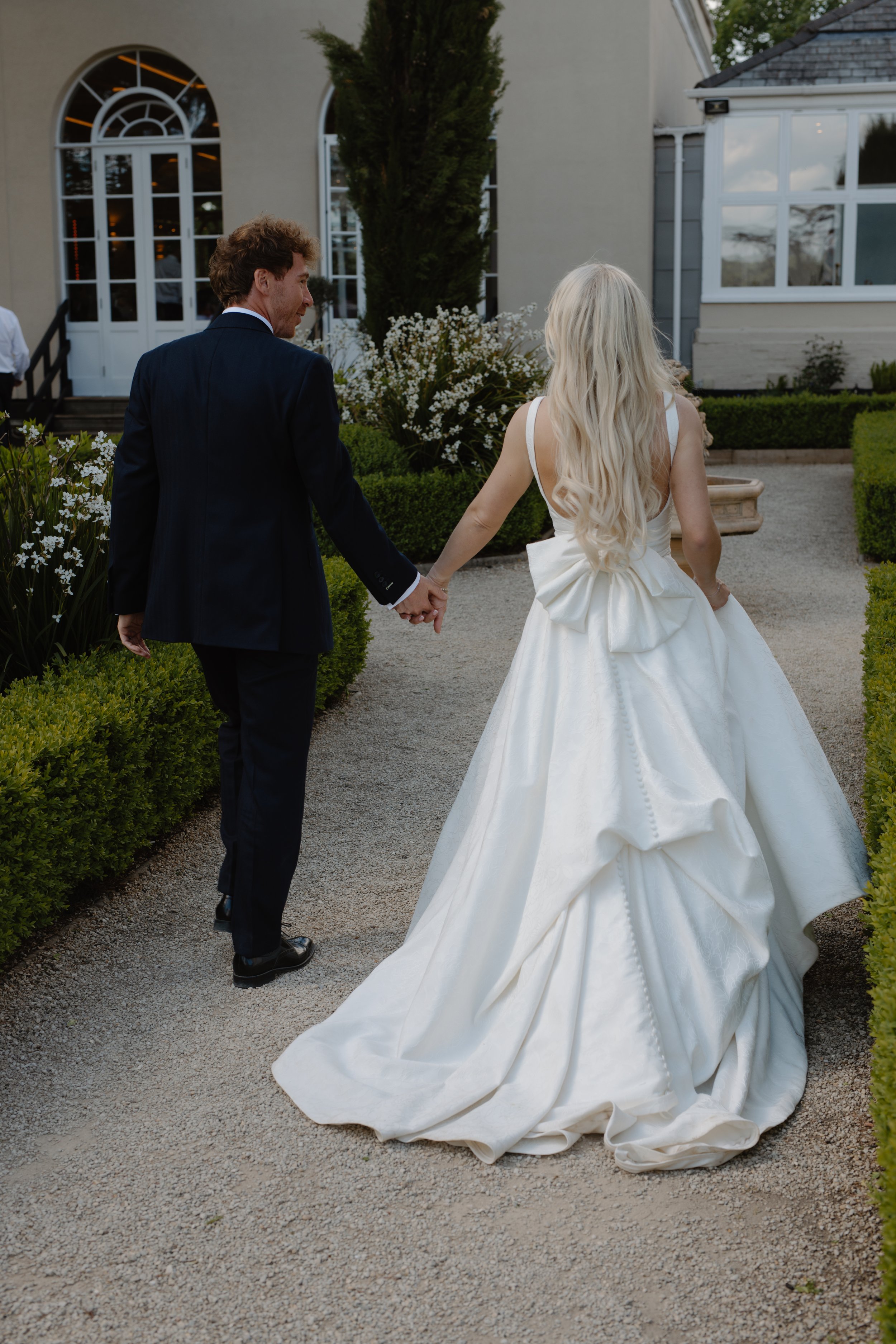 A bride and groom walking hand-in-hand on a garden path, with the bride wearing a white wedding gown with a large bow detail, and the groom in a dark suit, outside a white building with large windows.