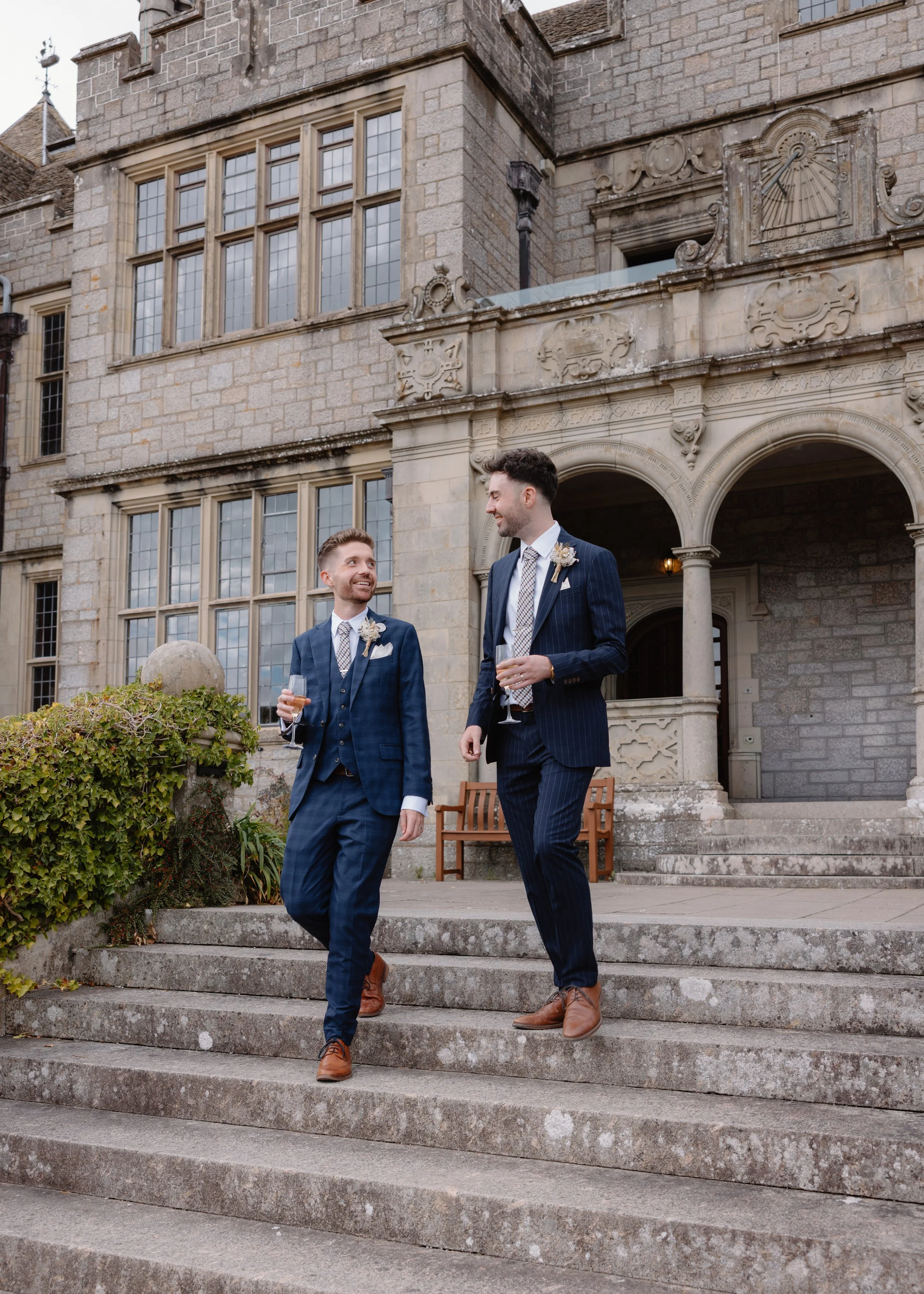 Two men dressed in suits, holding glasses of drinks, smiling and walking down stone steps outside a historic stone building with large windows and ornate architectural details.