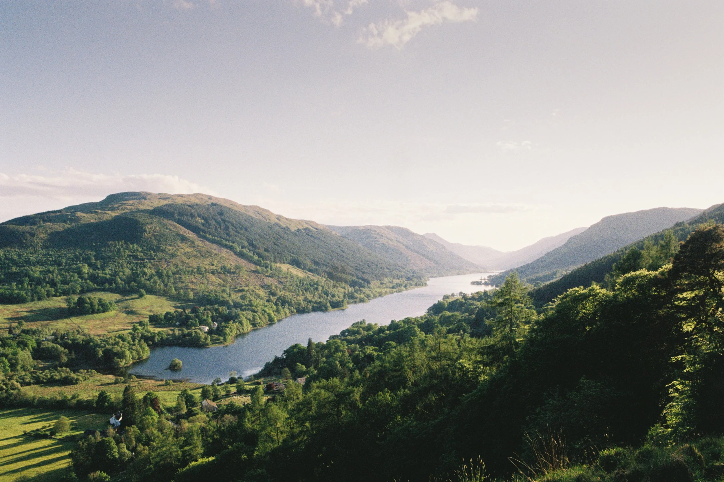 View of a lush green valley with a winding river, surrounded by tall mountains, under a partly cloudy sky.