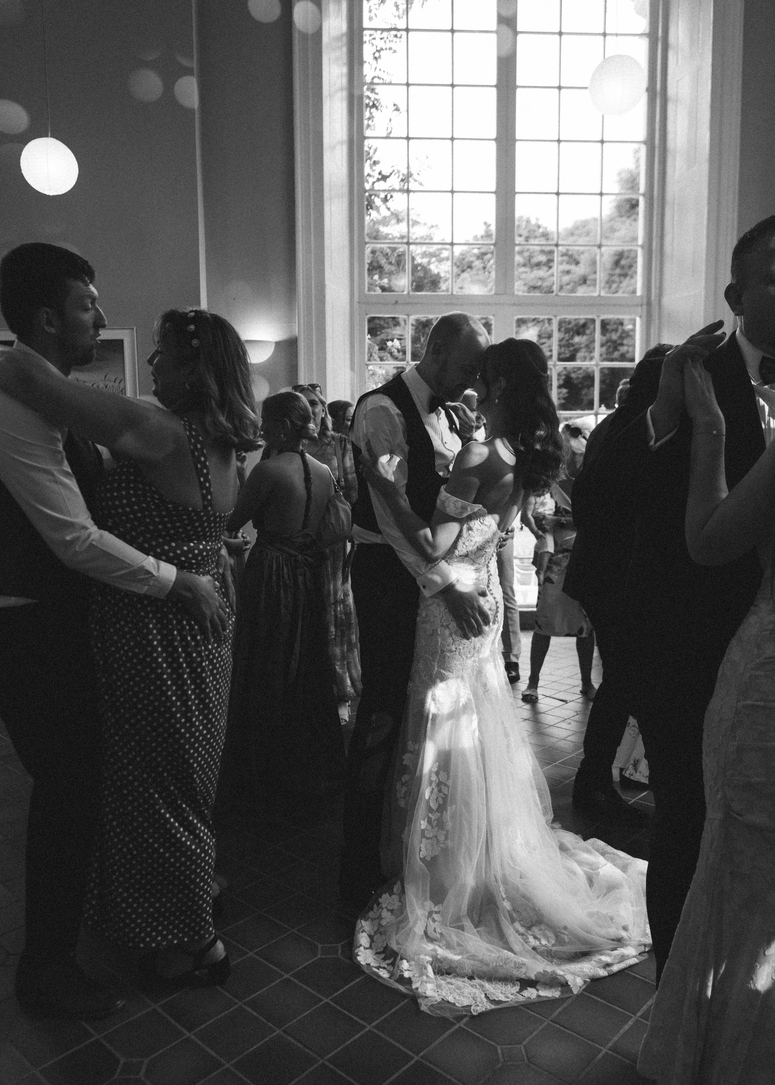 A black and white photo of a wedding reception with couples dancing, a bride and groom sharing a dance, and large windows in the background.