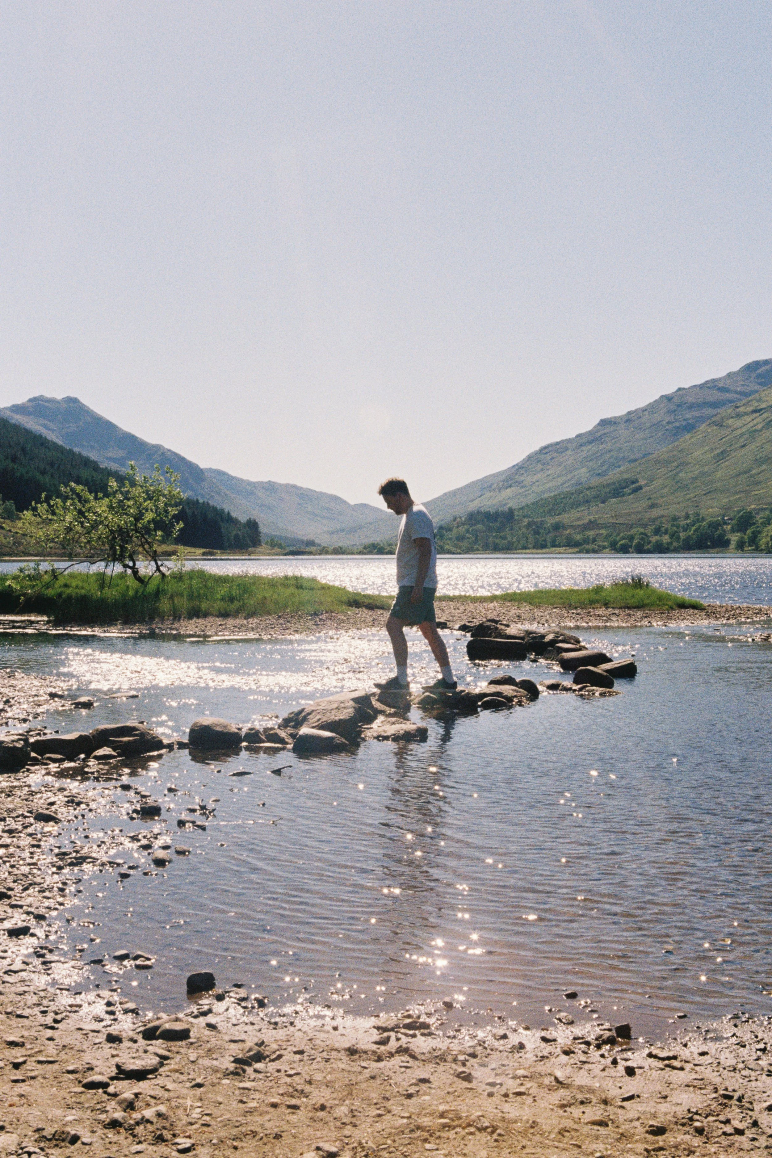 A person walking on rocks in a shallow river, with mountains and trees in the background under a clear sky.