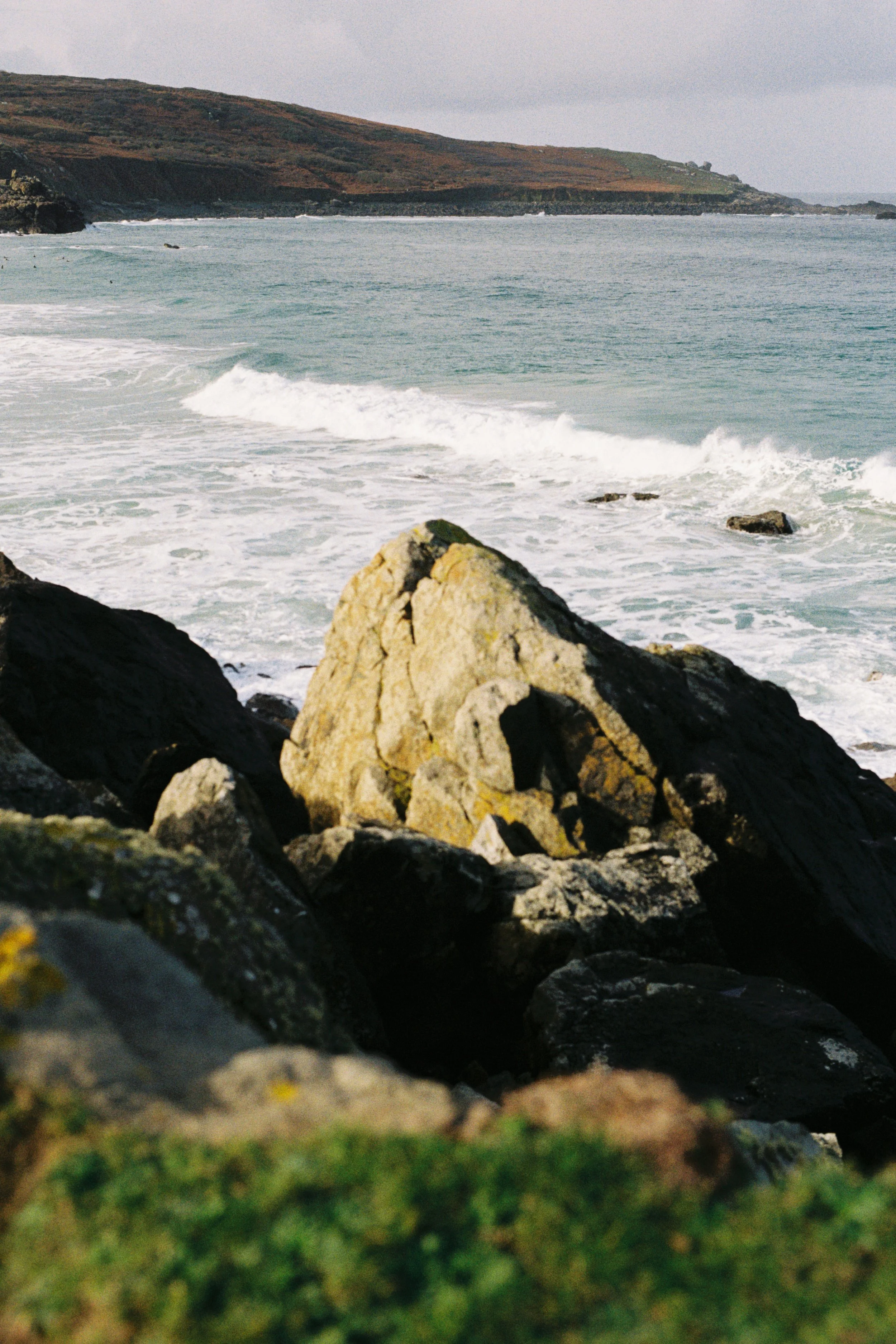 Rocks and moss along a beach overlooking the ocean with hills in the background.