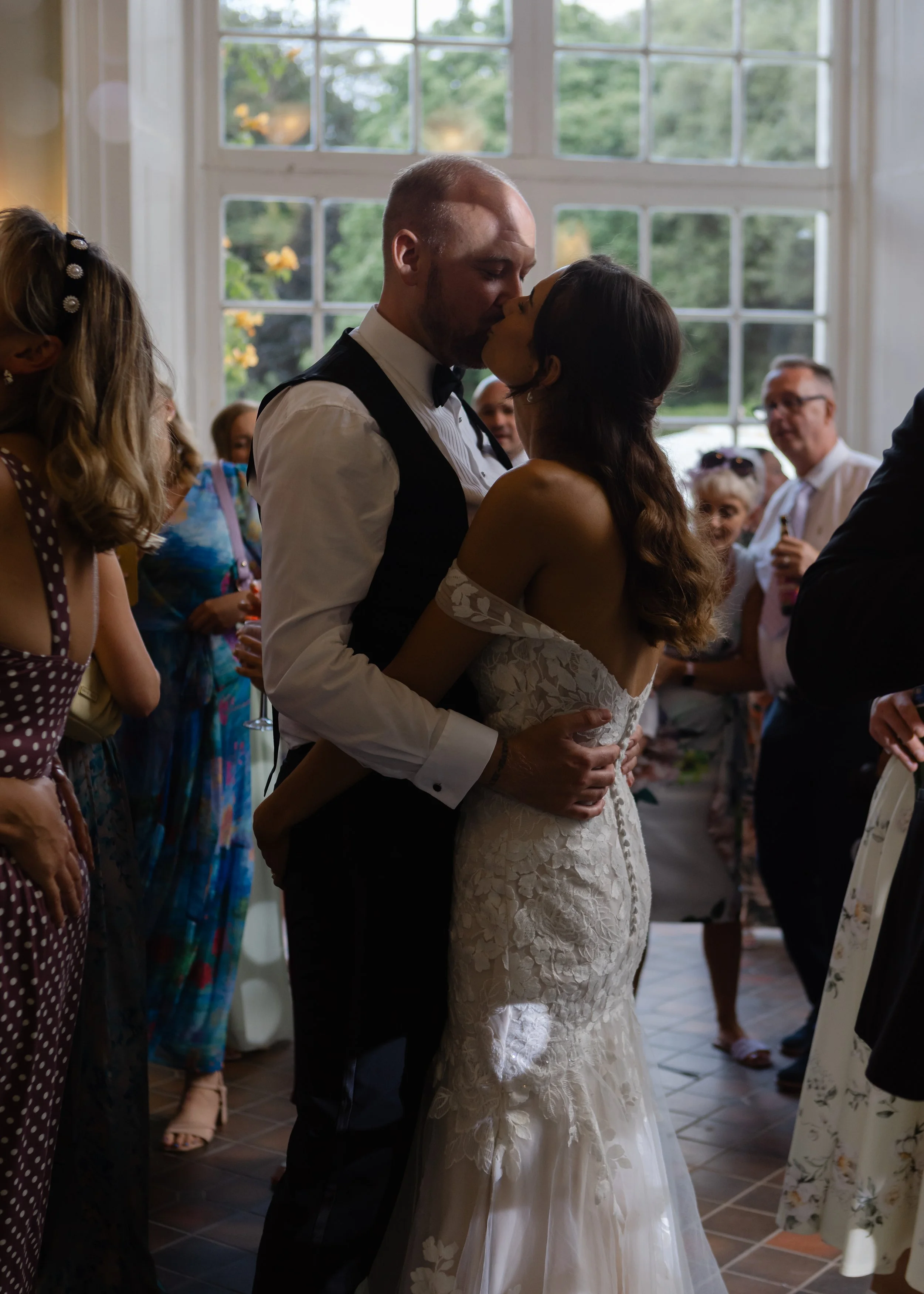 A bride and groom sharing a kiss at their wedding reception, surrounded by guests inside a well-lit room with large windows showing greenery outside.