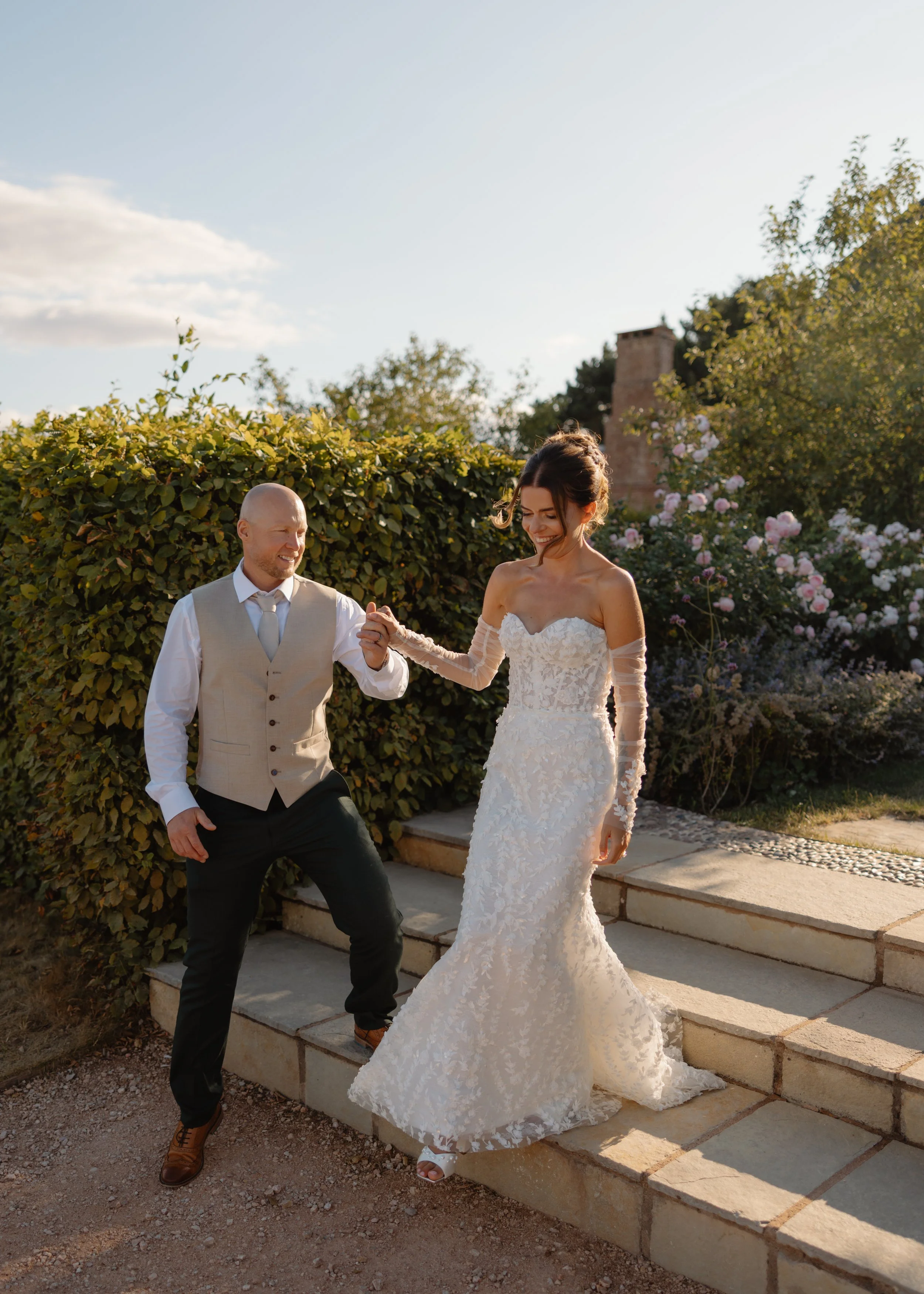 A bride and groom dancing on outdoor stone steps with greenery and pink flowers in the background, under a bright sky.