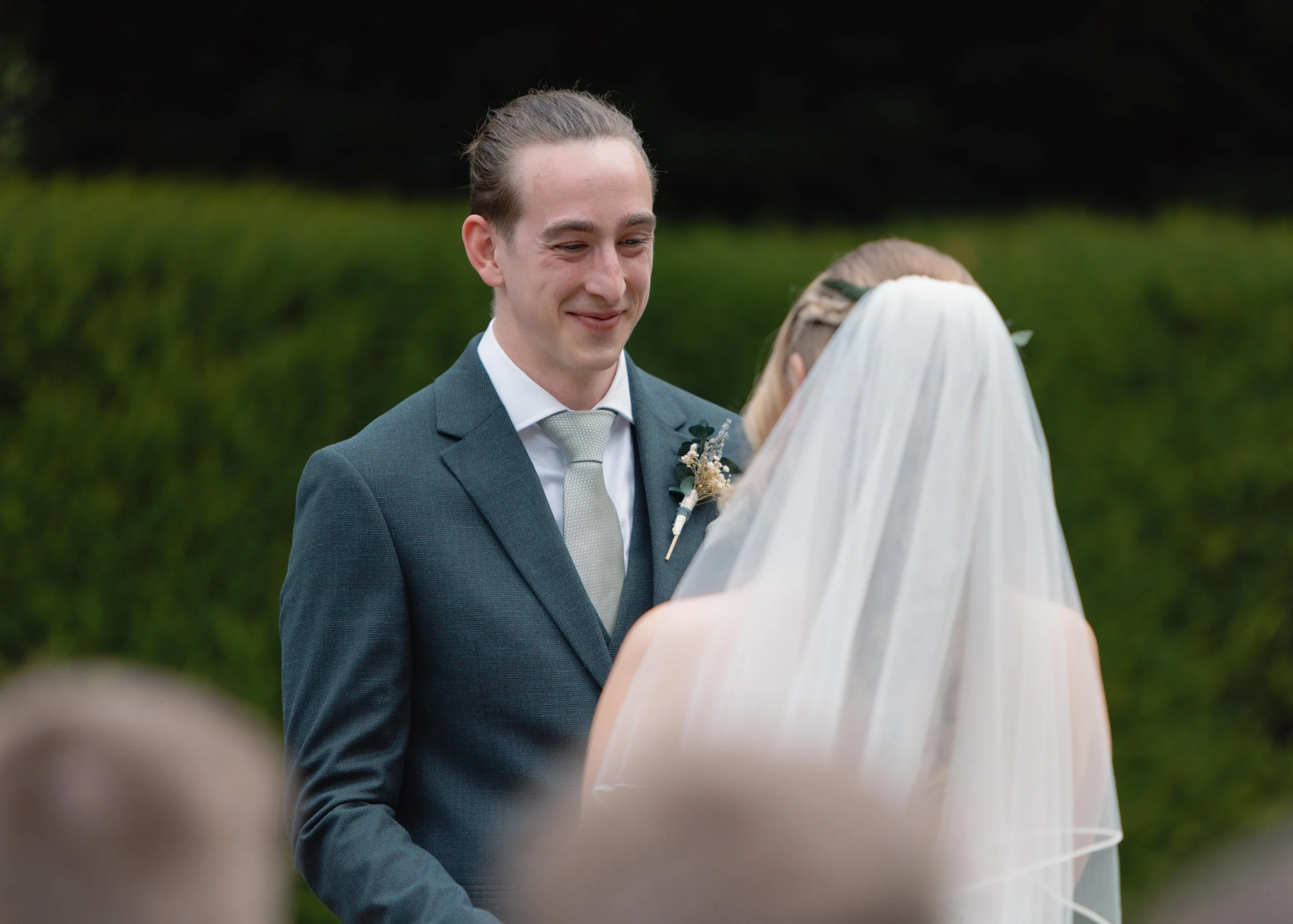 A groom smiling at a bride during a wedding ceremony outdoors with greenery in the background.