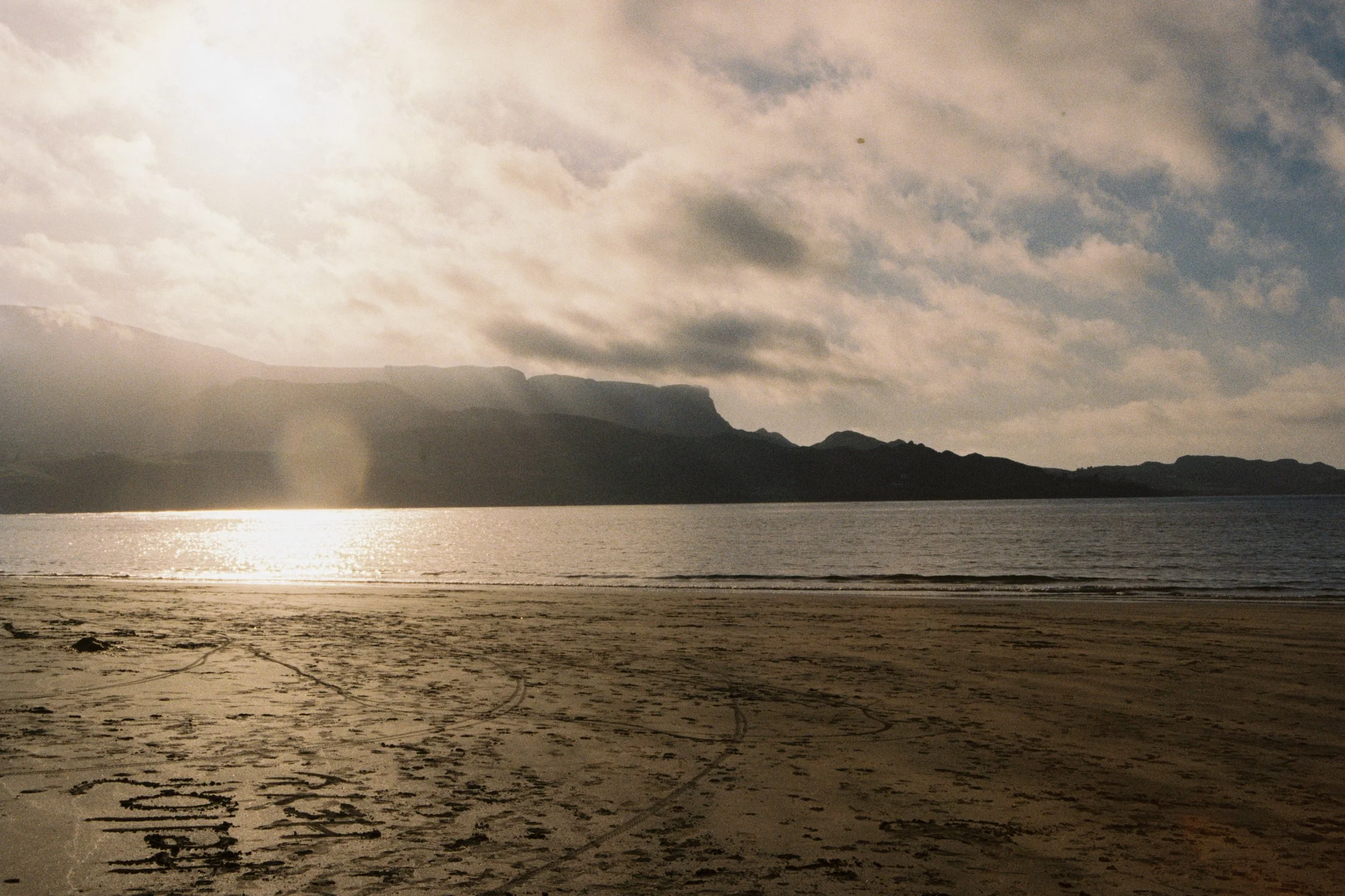 A serene beach scene with dark sand, calm water, mountains in the background, and a cloudy sky with sunlight breaking through.