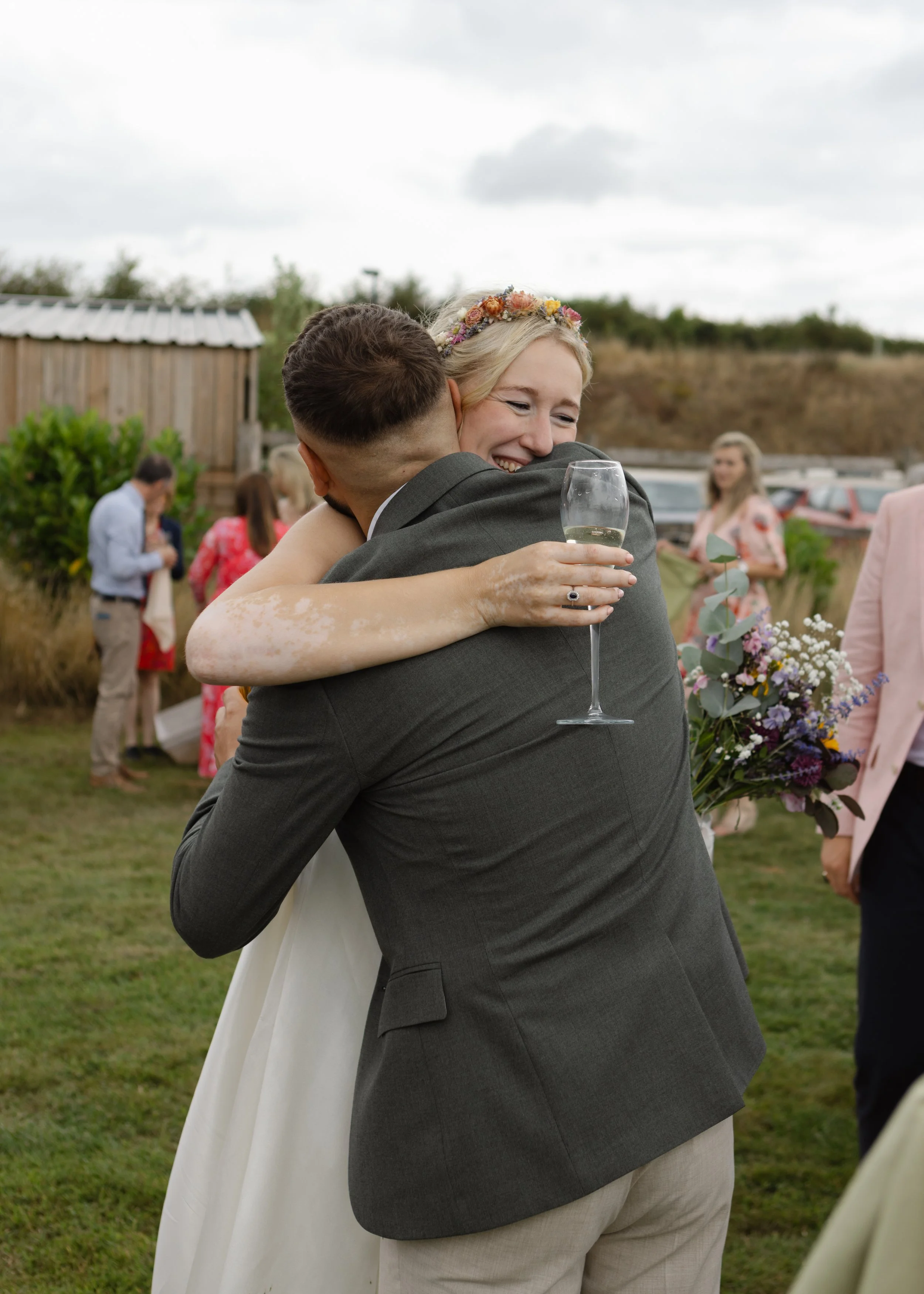 Two people hugging during an outdoor wedding celebration, one holding a glass of champagne.