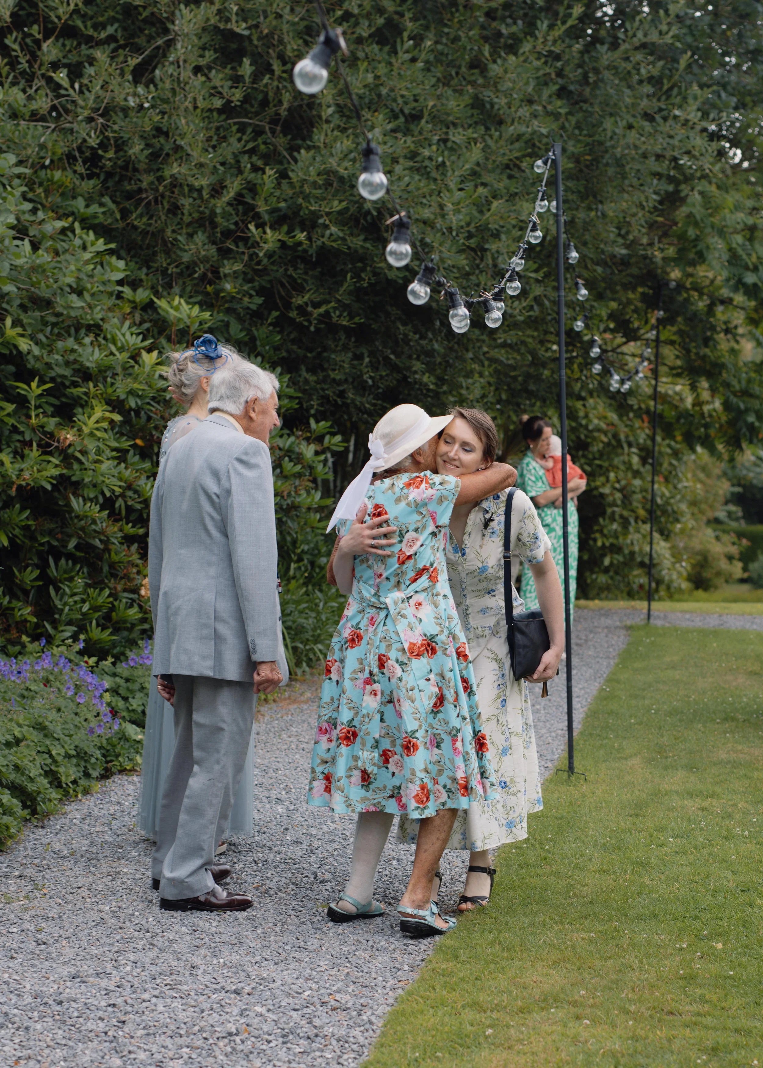 Four people hugging and chatting outdoors at a garden party decorated with string lights, with greenery and a gravel pathway.
