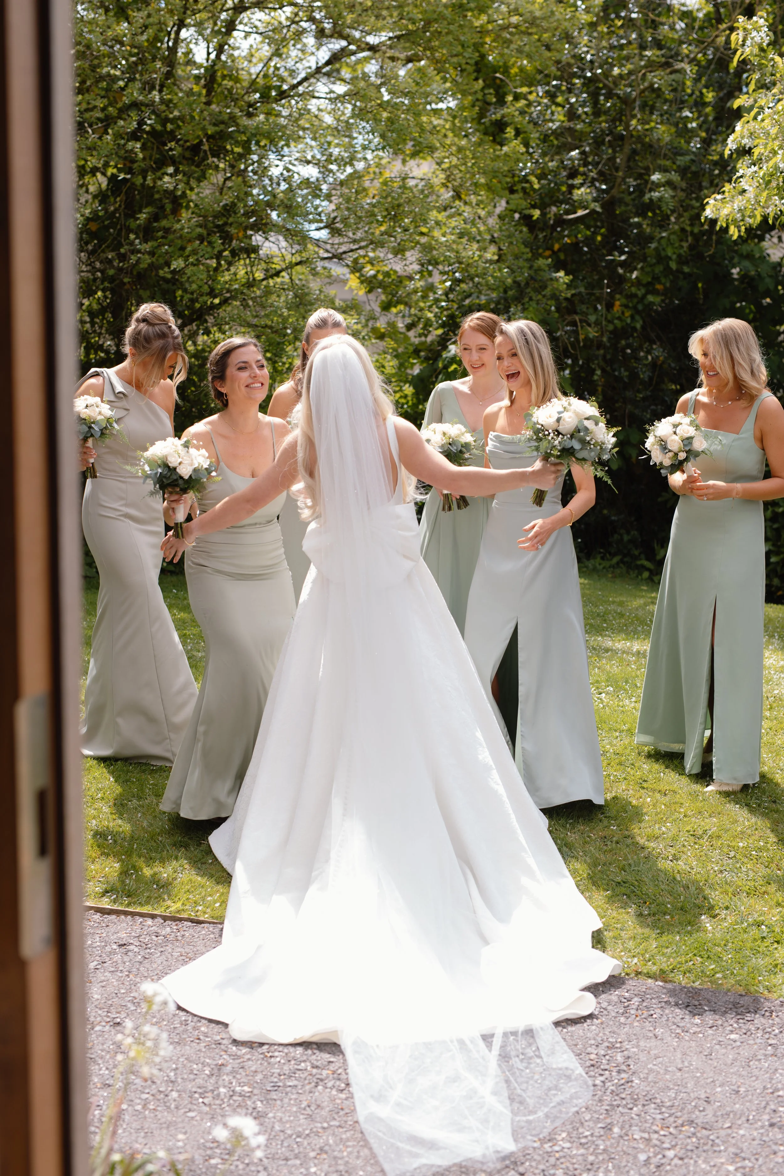 Bride in wedding dress and veil greeting five bridesmaids holding bouquets in a garden with trees in the background.