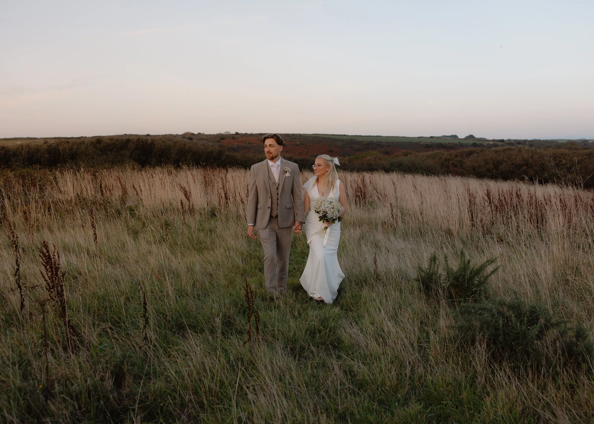 A couple walking hand-in-hand through a grassy field at sunset, the bride in a white wedding dress holding a bouquet and the groom in a light gray suit with a vest.
