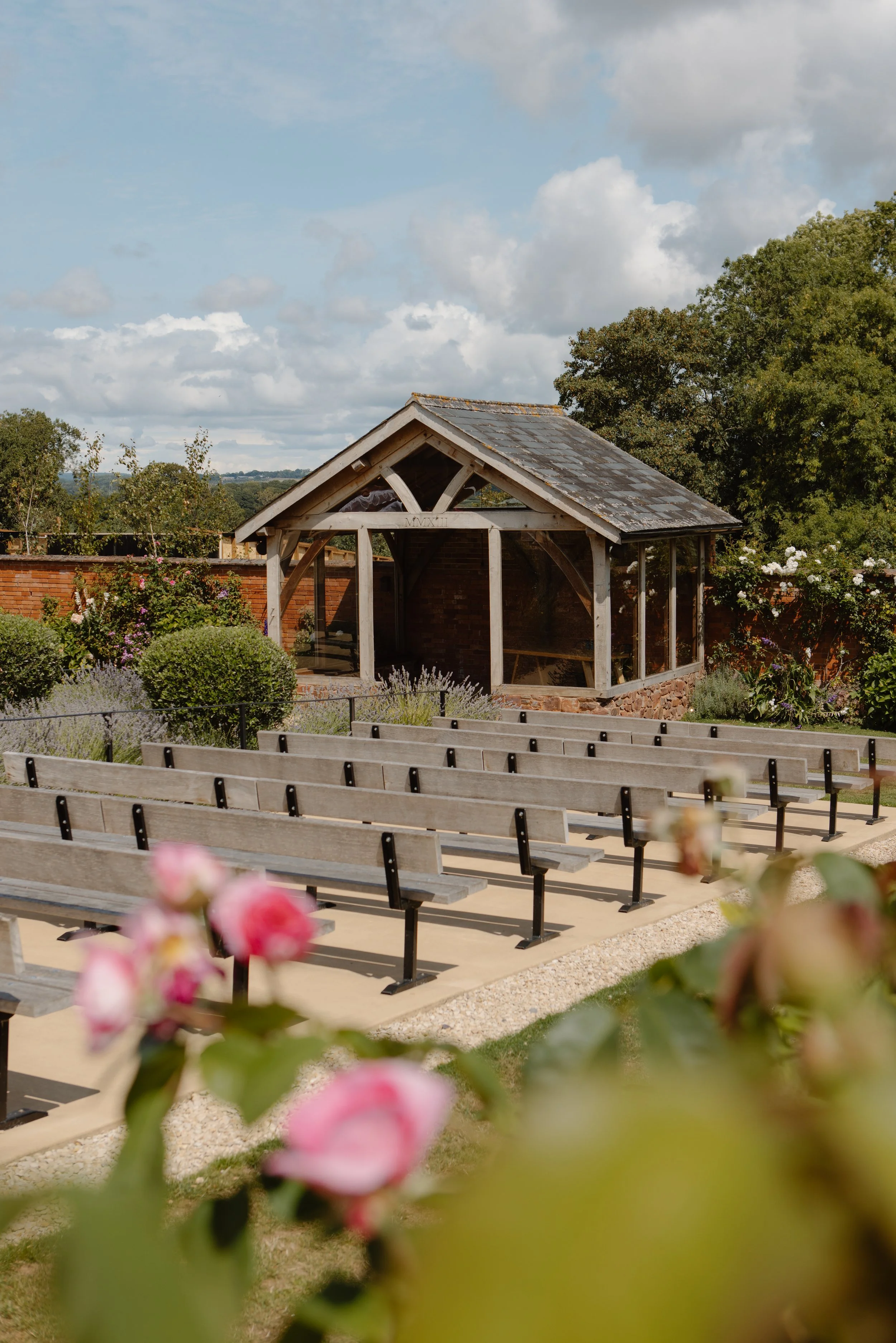 An outdoor scene with wooden benches facing a small wooden and glass structure, surrounded by flowering bushes and trees under a partly cloudy sky.