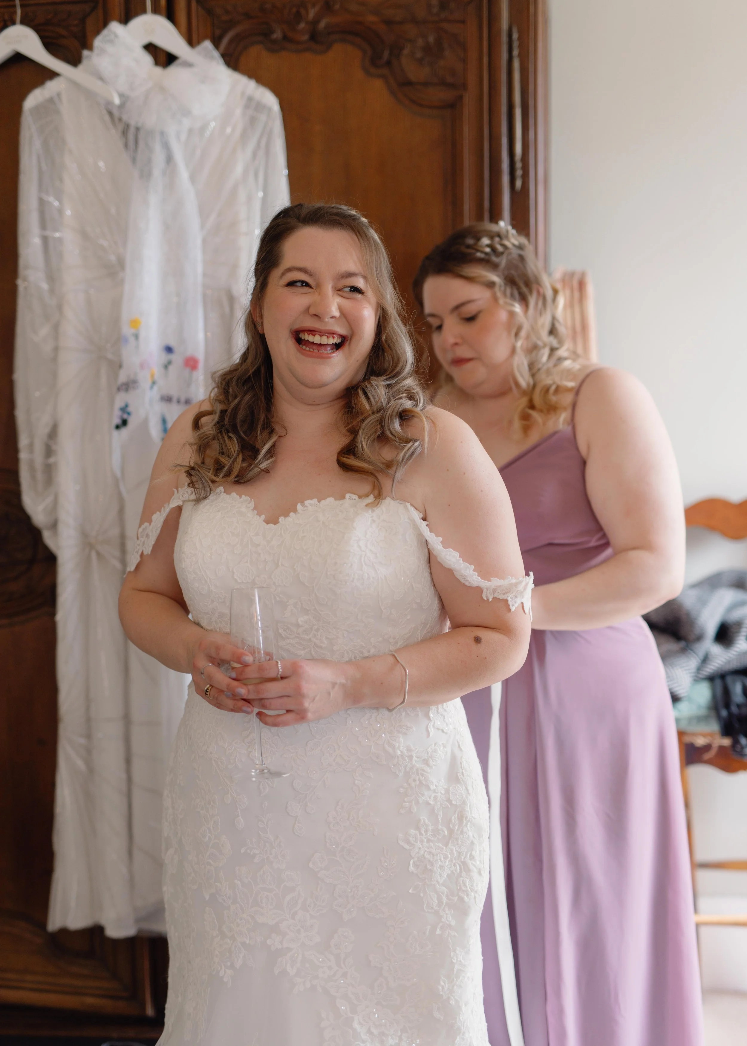 Bride in a white lace wedding dress holding a glass, smiling joyfully, in a room with a woman in lavender dress preparing behind her, and a wedding dress hanging on a wooden wardrobe in the background.
