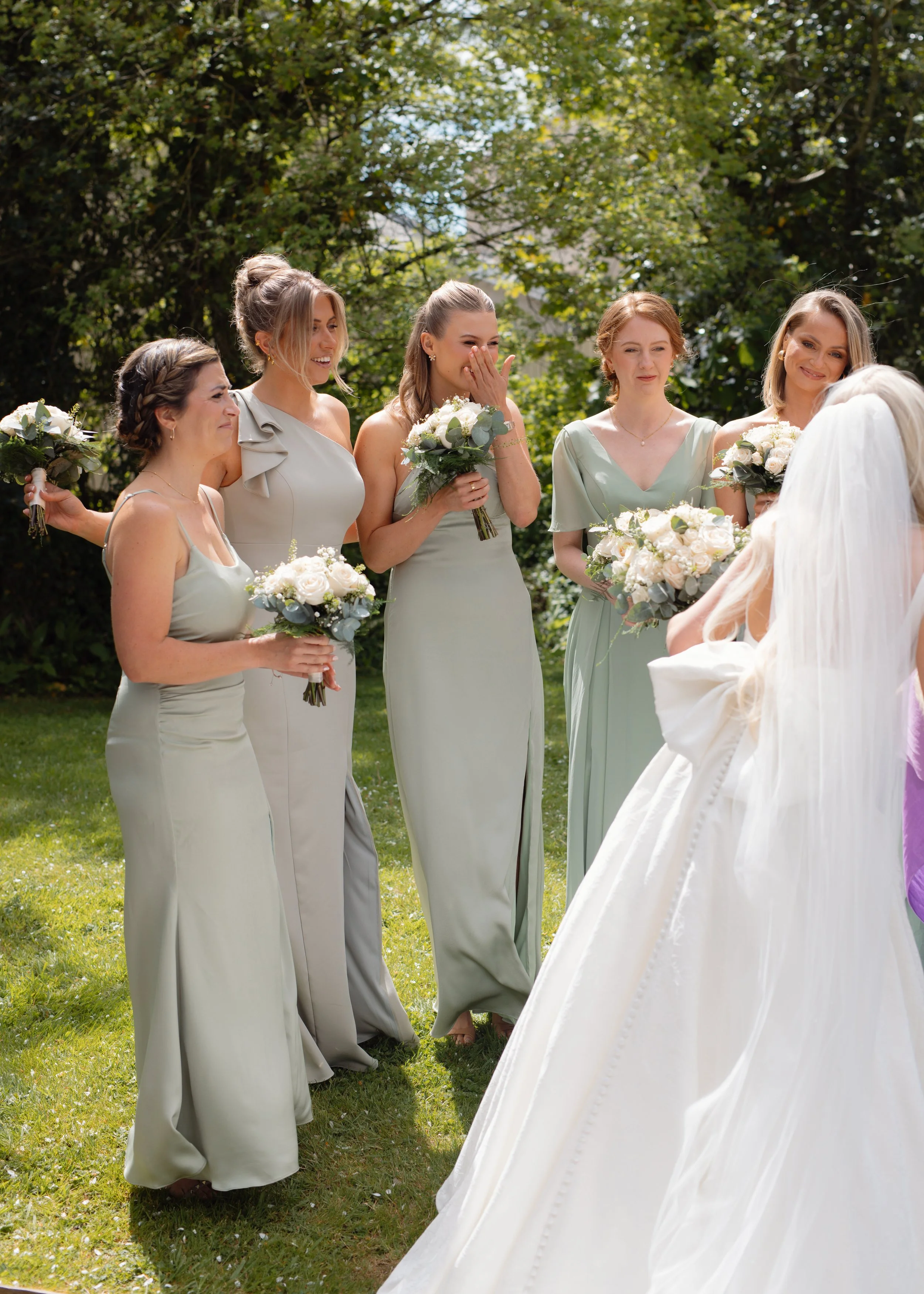 Bride and bridesmaids during a wedding ceremony outdoors, with the bridesmaids holding bouquets of white and green flowers, and the bride in a white wedding dress with a veil.