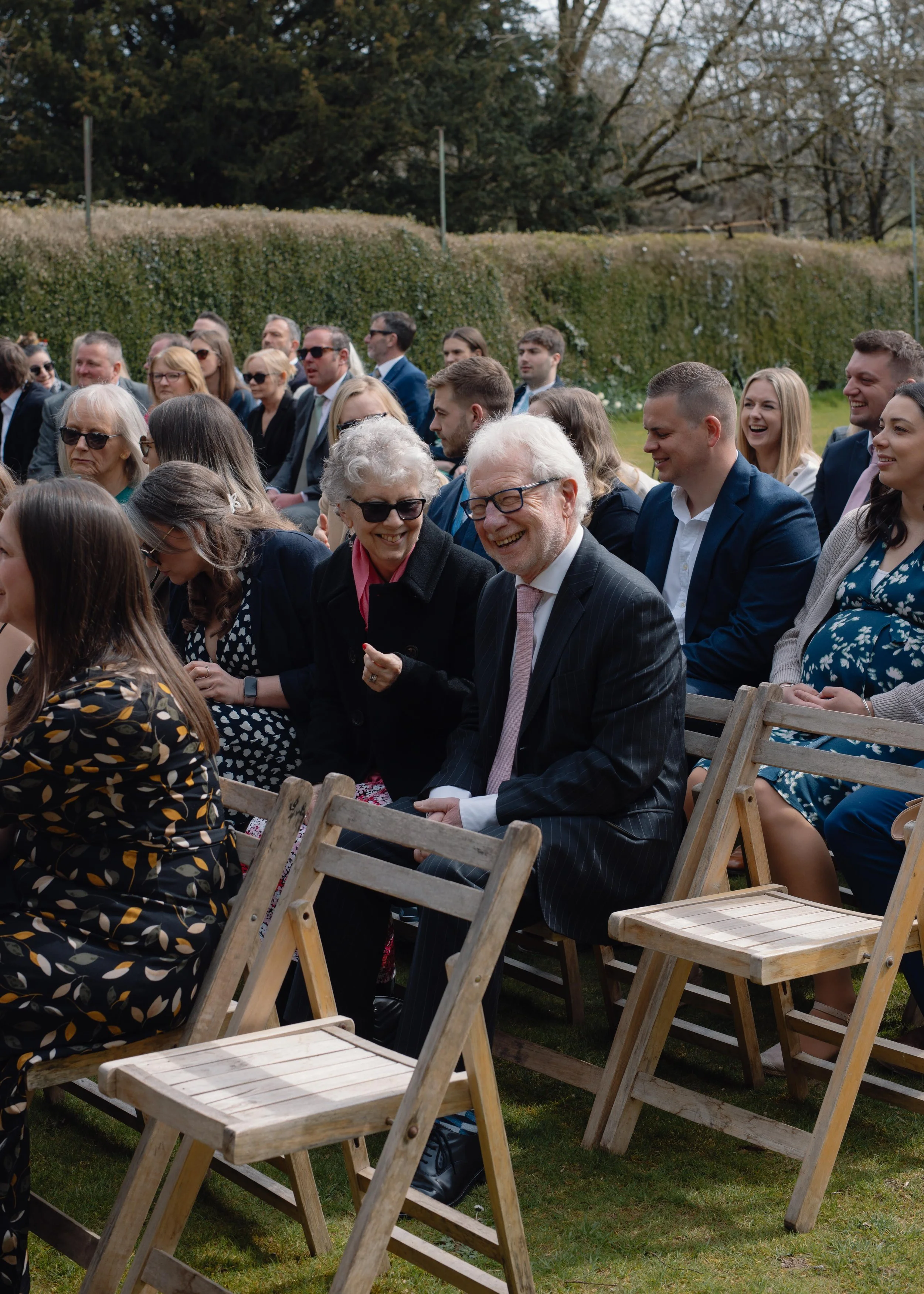 A group of people dressed in formal attire sitting outdoors on wooden chairs at a joyful event, likely wedding or celebration, with trees and bushes in the background.