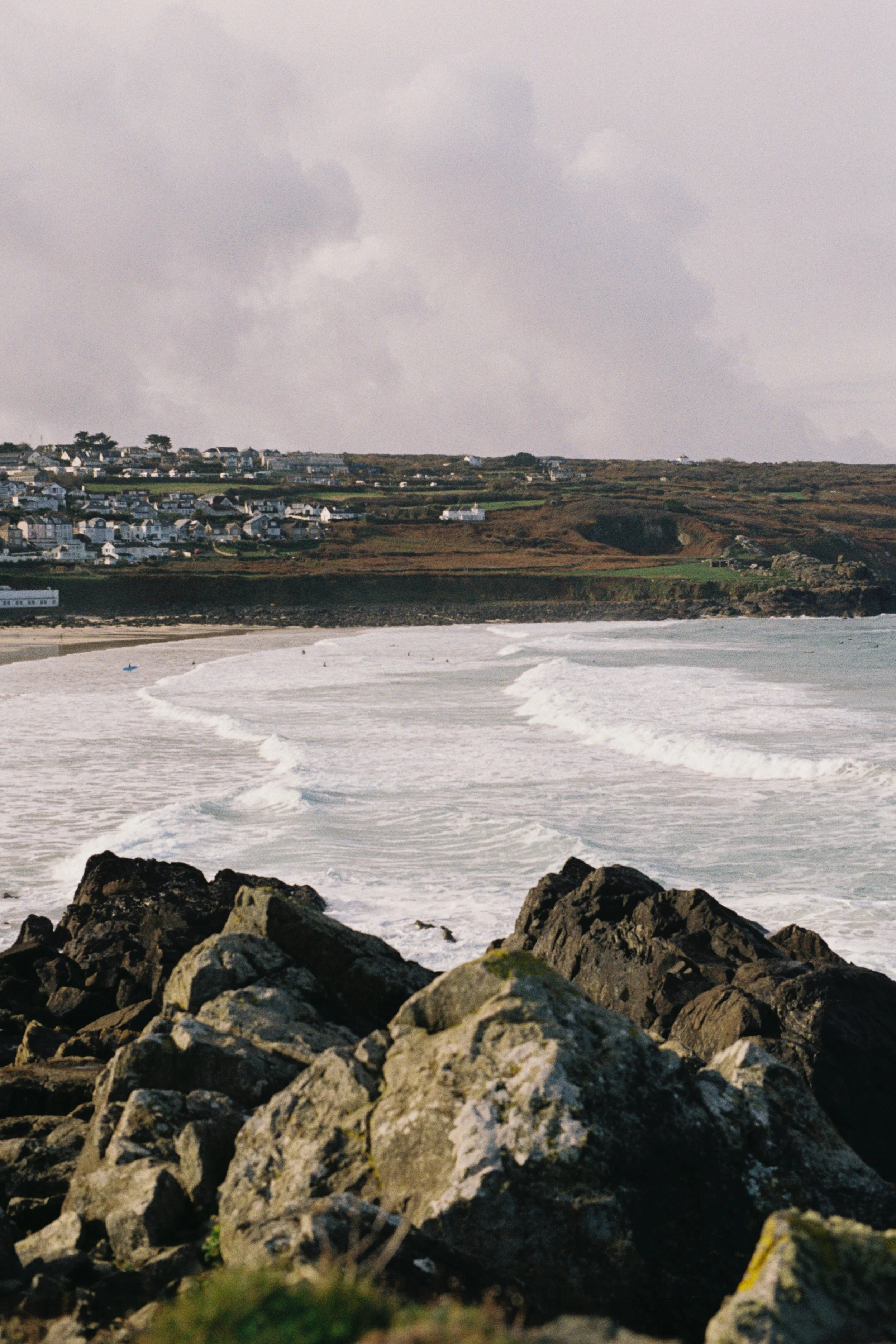 Rocky shoreline in the foreground with ocean waves and a distant hillside with houses, under a cloudy sky.