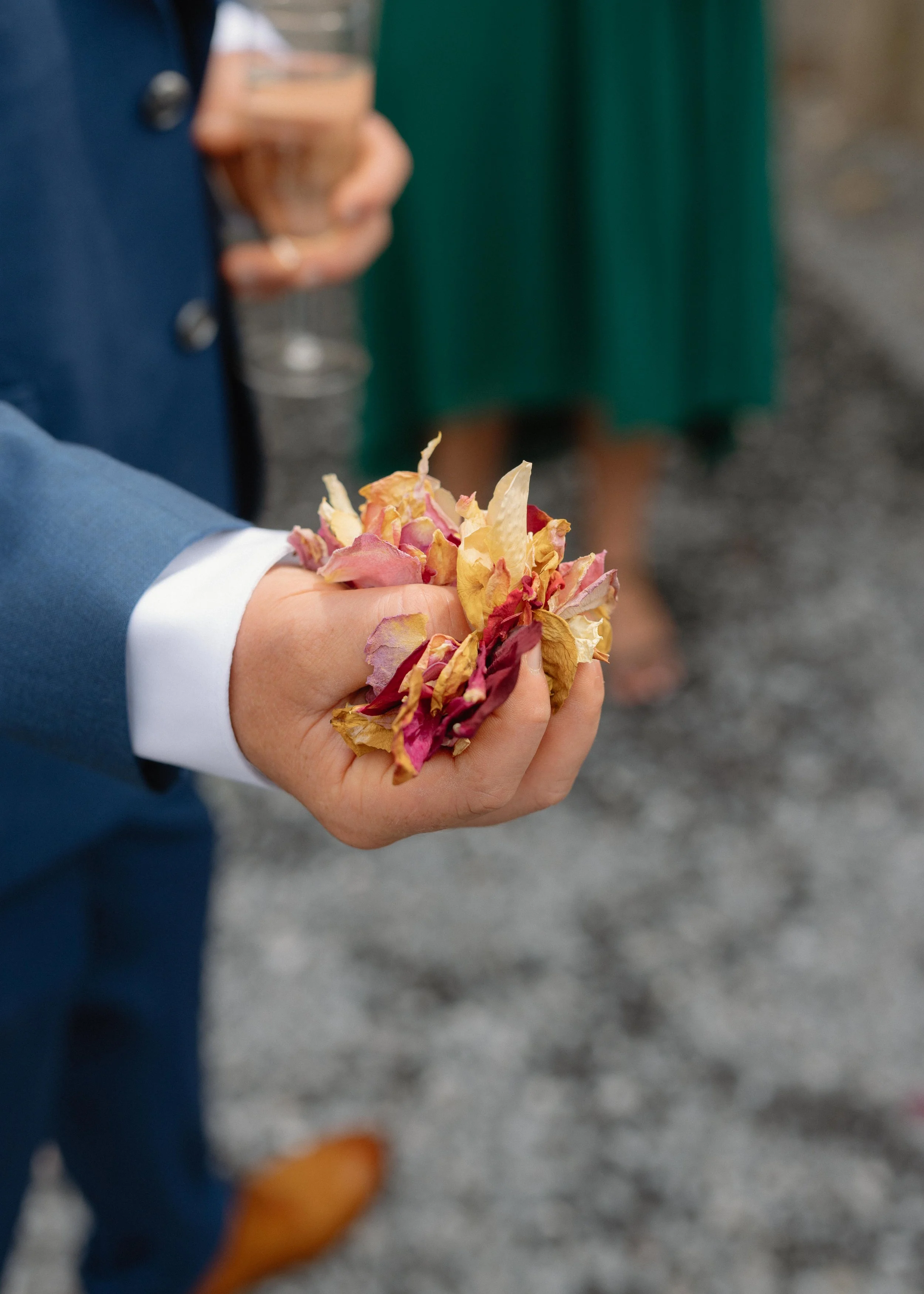 Close-up of a person in a blue suit holding colorful dried flower petals in their hand, with a green dress and a glass of champagne in the background.