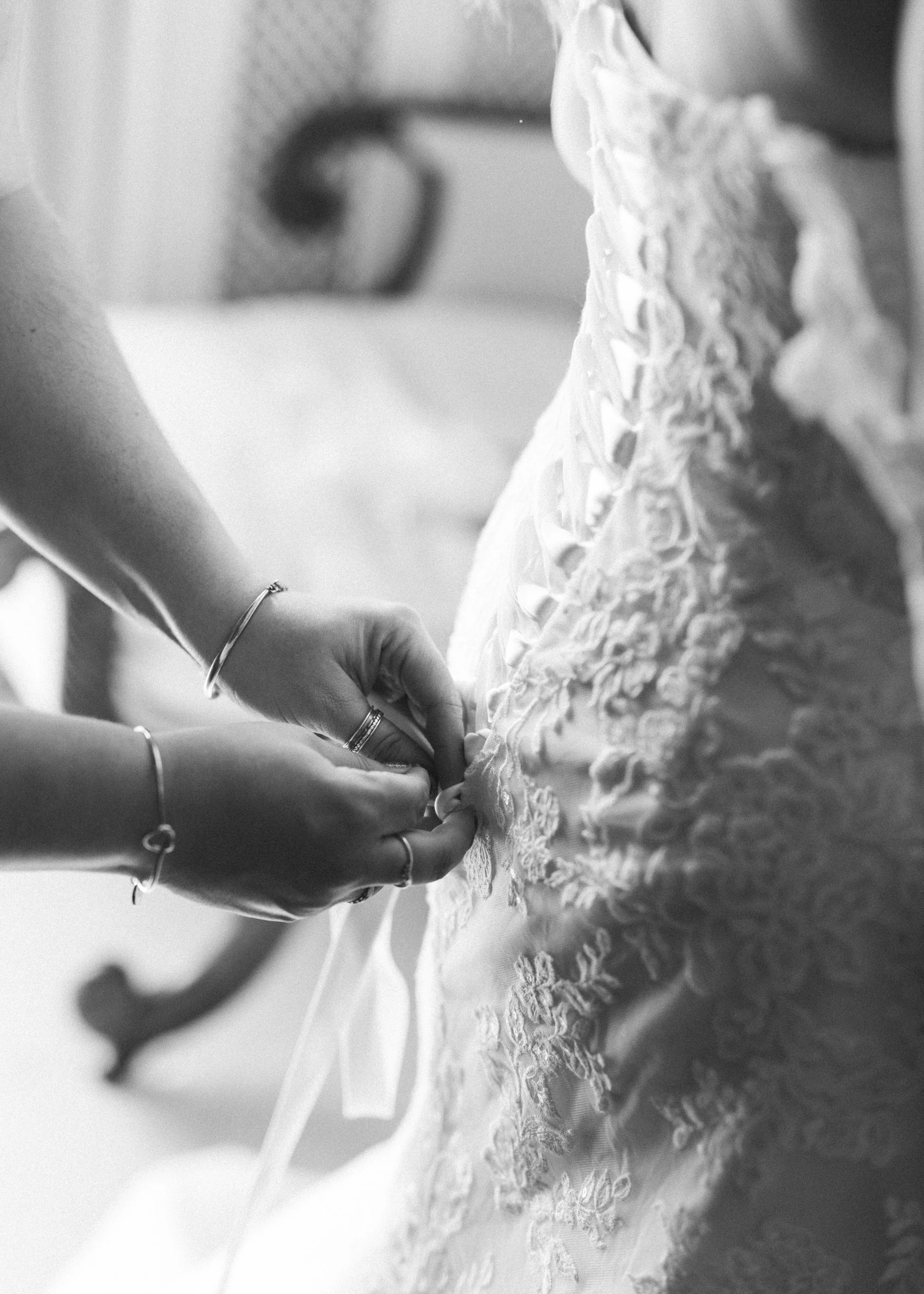 A woman fastening the back of a wedding dress with lace and ruffles, captured in black and white.