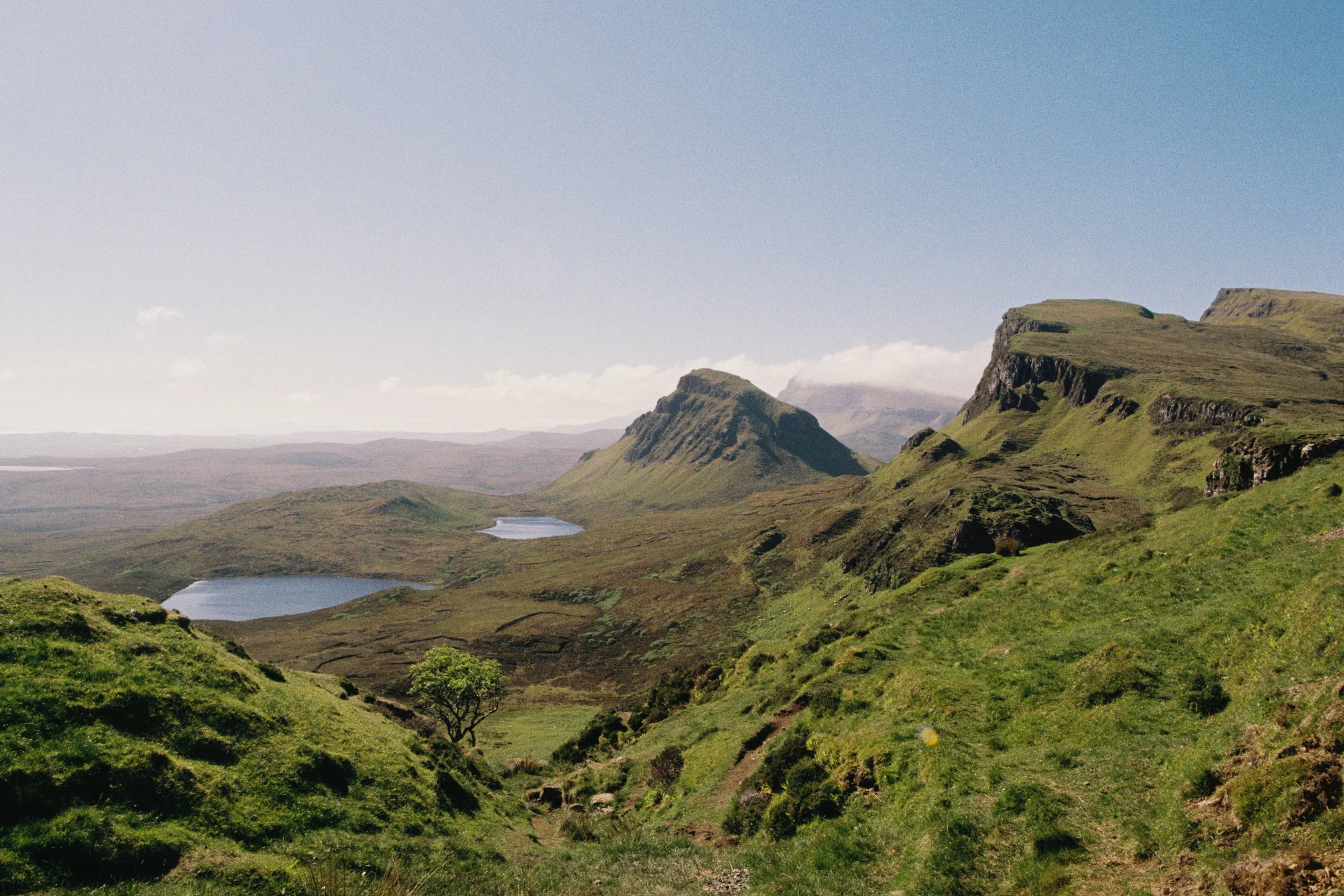 A landscape of green rolling hills with small lakes, rocky formations, and a partly cloudy sky.