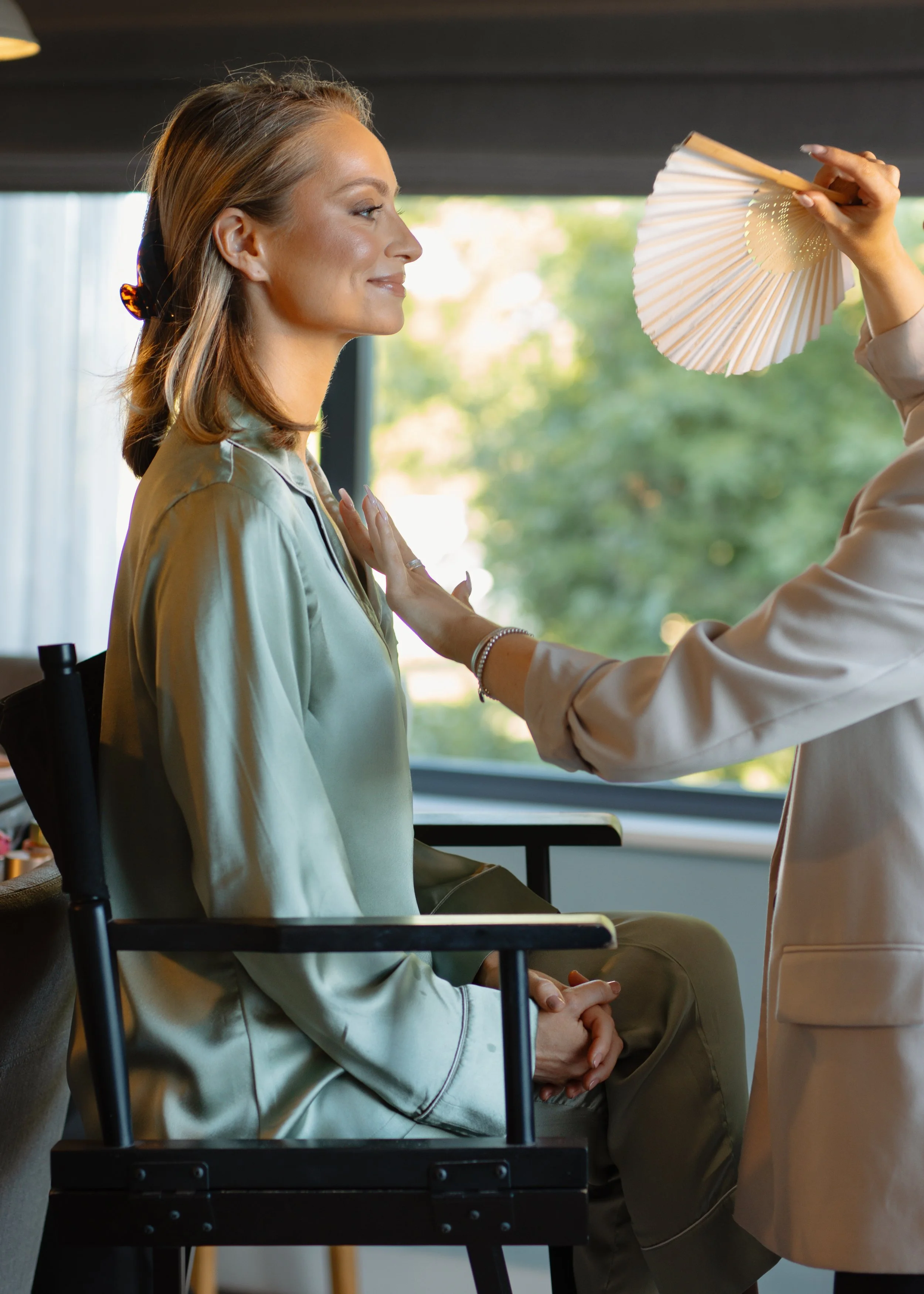 A woman with light brown hair and a satin robe sitting on a chair with her eyes closed as a makeup artist applies makeup with a fan brush, indoors with natural light coming through large windows.