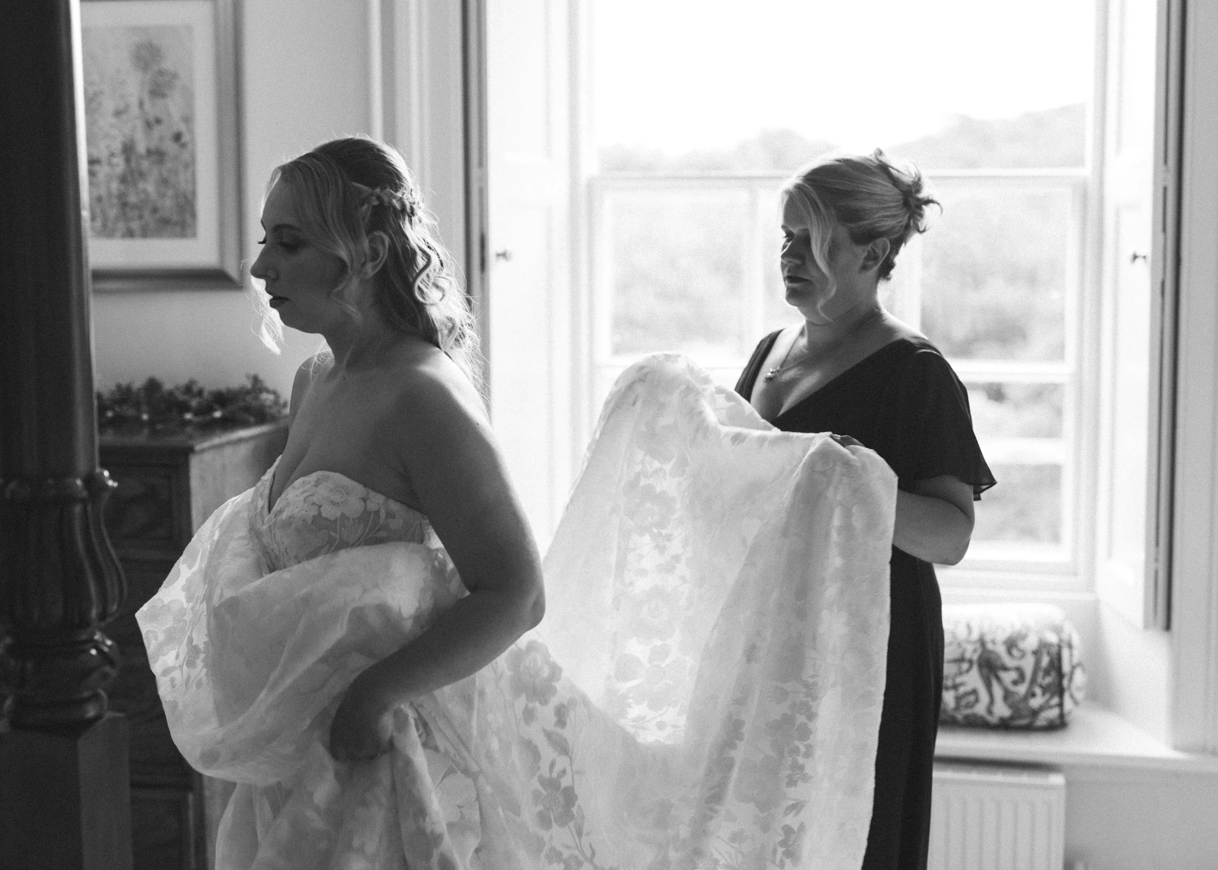 A woman in a strapless wedding dress stands in the foreground, with her eyes closed. Another woman behind her holds the train of the wedding dress. Both women are indoors near a window, in black and white.