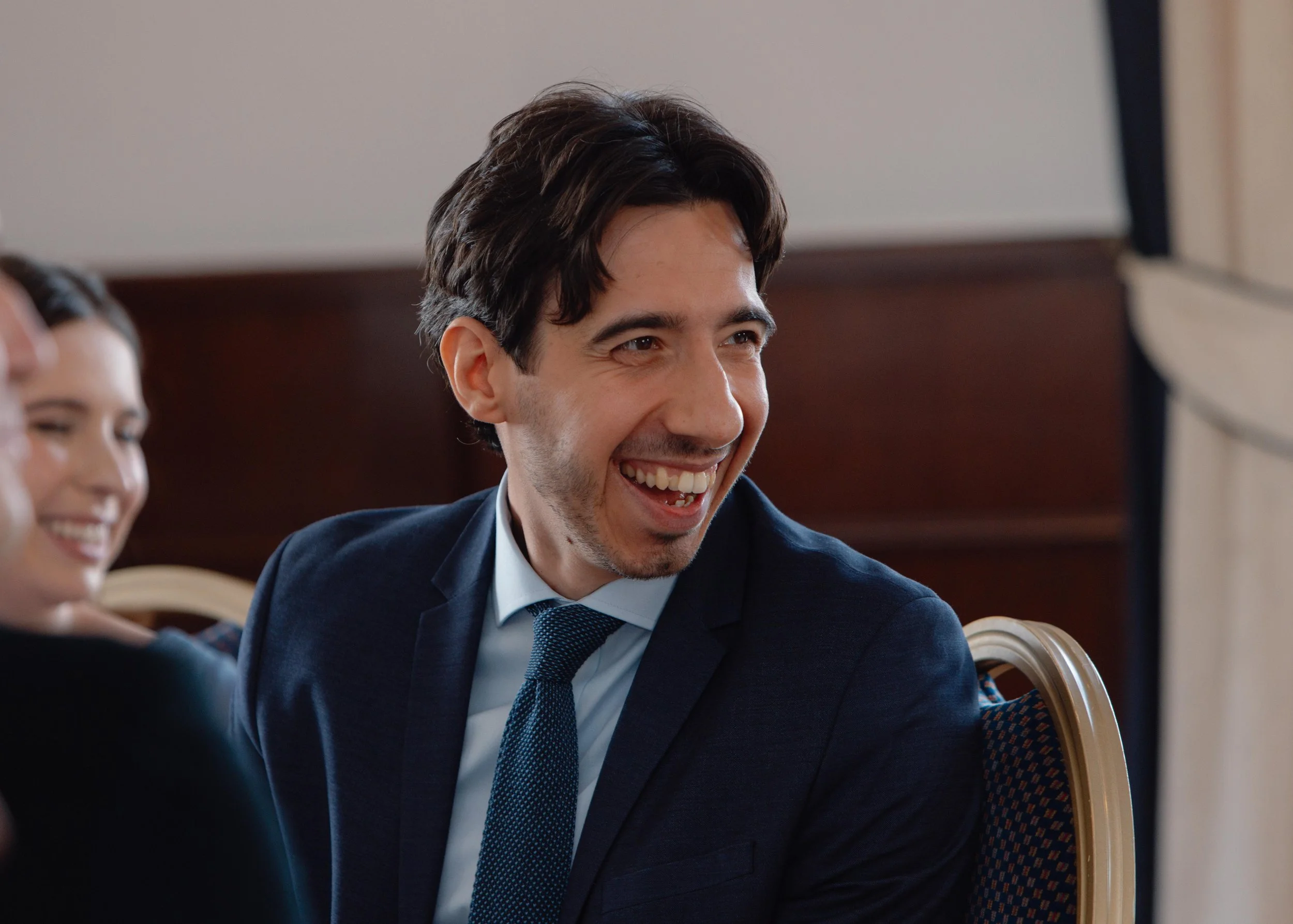 A man in a suit laughing and smiling at a formal event, with a woman beside him also smiling.