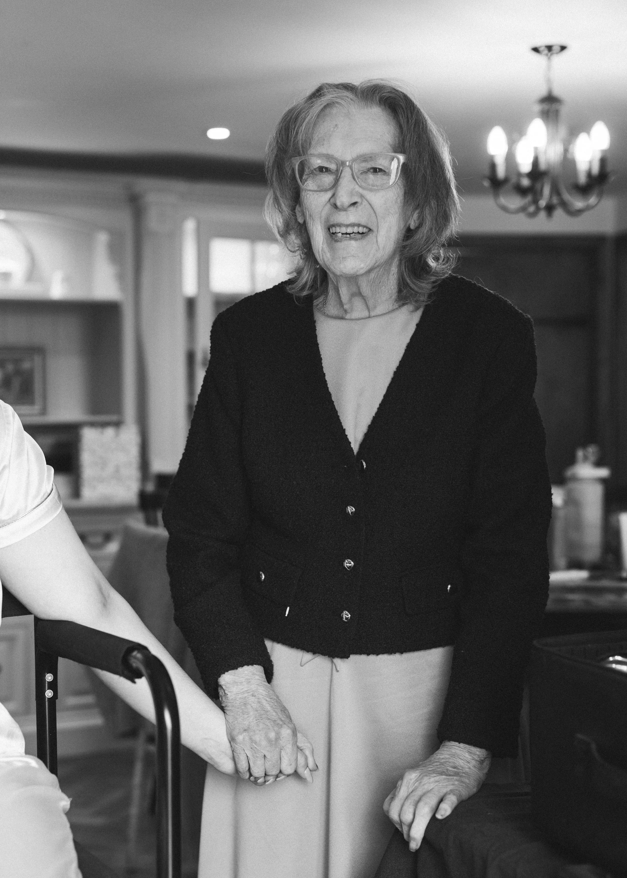 An elderly woman with glasses, wearing a black cardigan and light-colored pants, is holding hands with a person partly visible on the left side of the image in a home setting.
