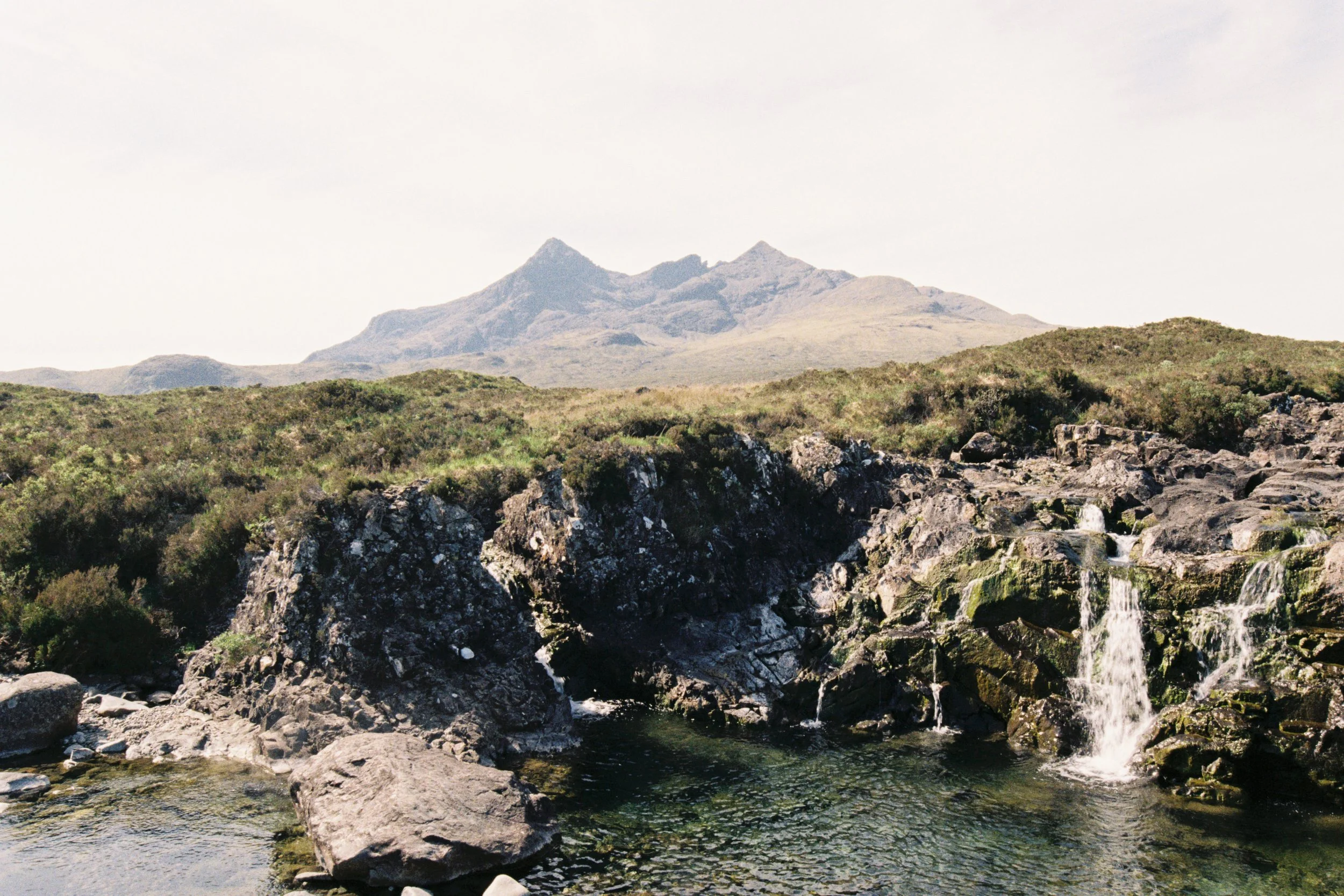 A mountain range in the background, with a river flowing over rocks and boulders in the foreground.