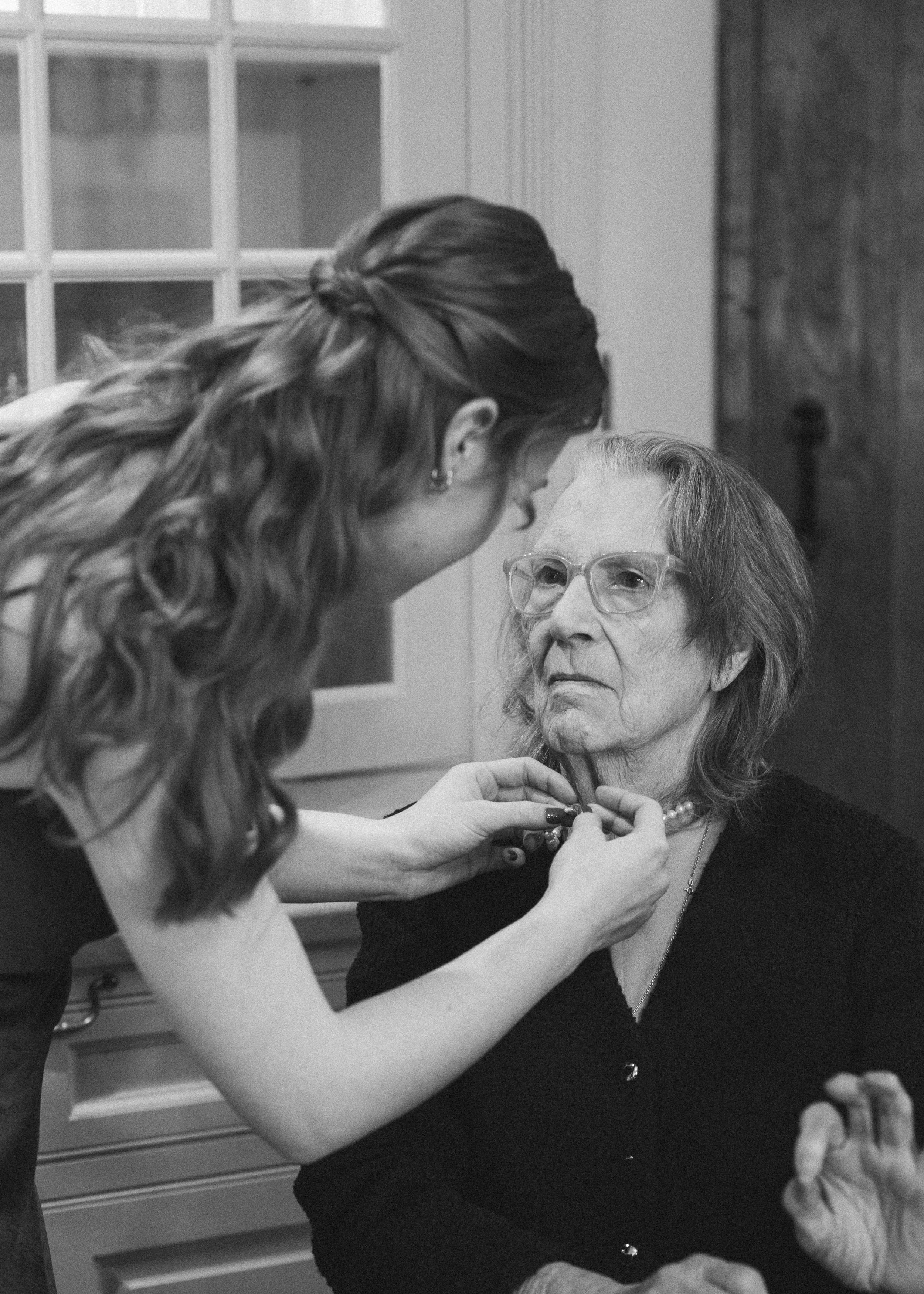 A young woman with long, wavy hair and earrings is adjusting the necklace of an elderly woman with glasses, shoulder-length hair, and a serious expression, seated indoors.