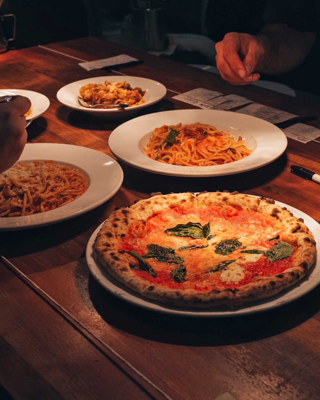 A wood-fired pizza and several pasta plates served on a wooden table.
