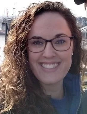 Close-up of a woman with curly hair, glasses, and a broad smile outdoors.