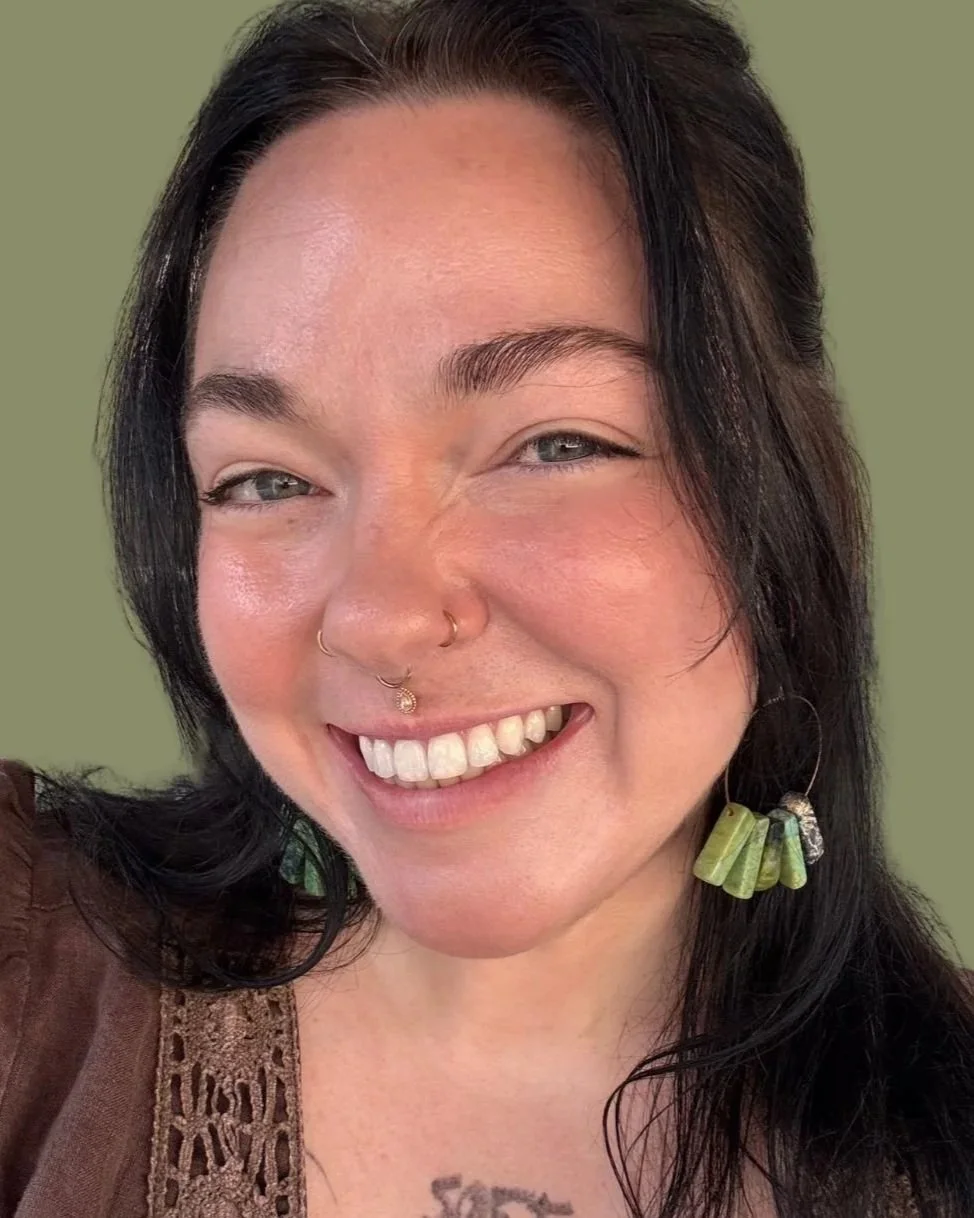 A young woman smiling with black hair, green eyes, multiple facial piercings, and wearing large earrings. She is wearing a brown top with lace detail.