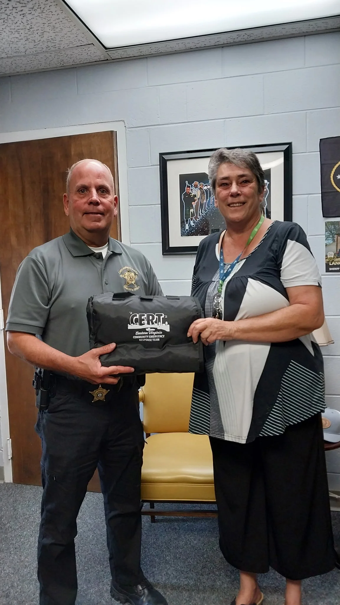 A police officer and a woman standing indoors, holding a CERT Emergency Response Team kit. The police officer is wearing a gray uniform with badge insignia, and the woman is dressed in a black and white outfit with a lanyard around her neck.