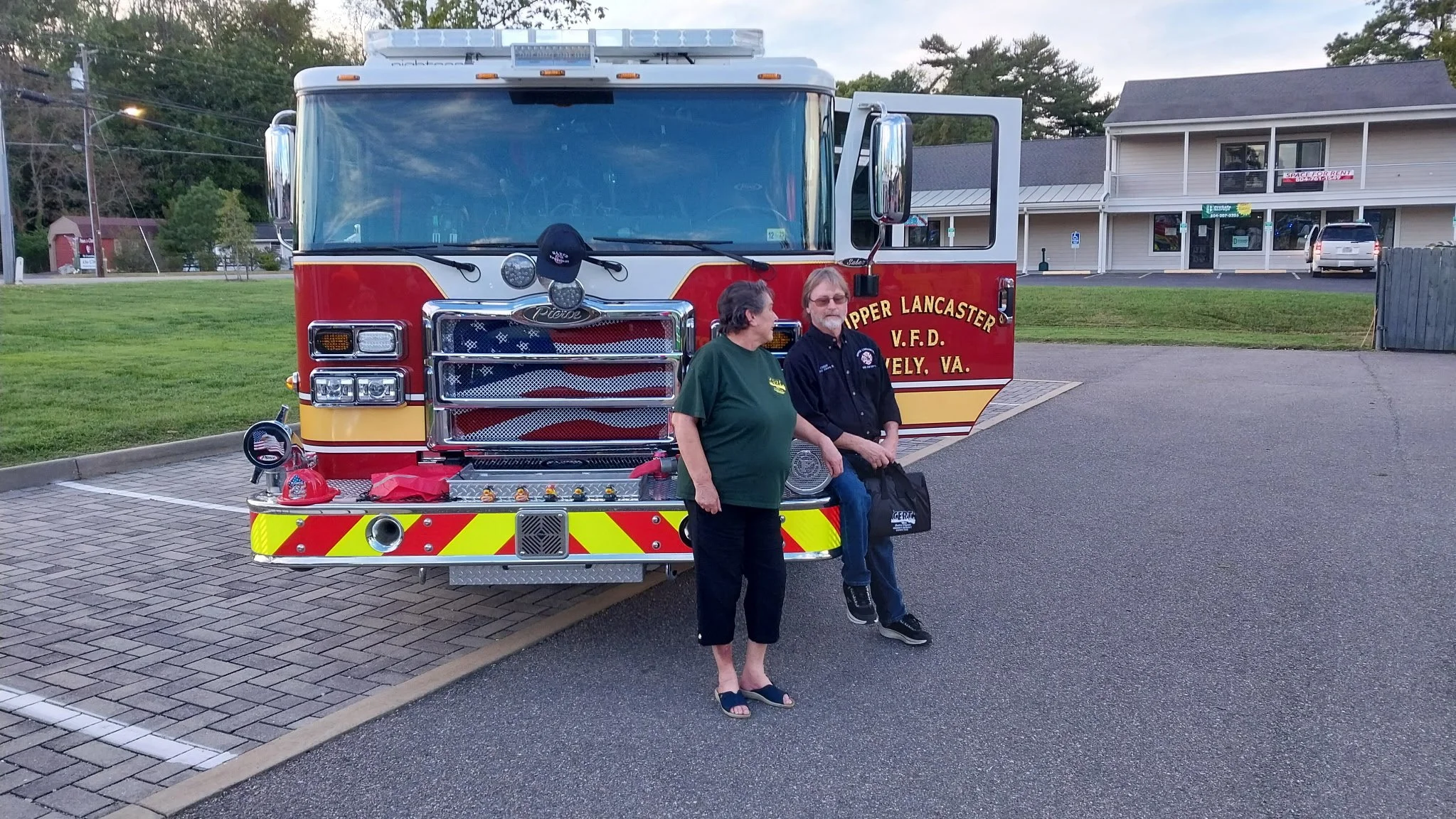 Two people standing and sitting in front of a fire truck with 'Upper Lancaster V.F.D., Kewlwy, VA.' written on the side, parked on a paved area with a grassy background.