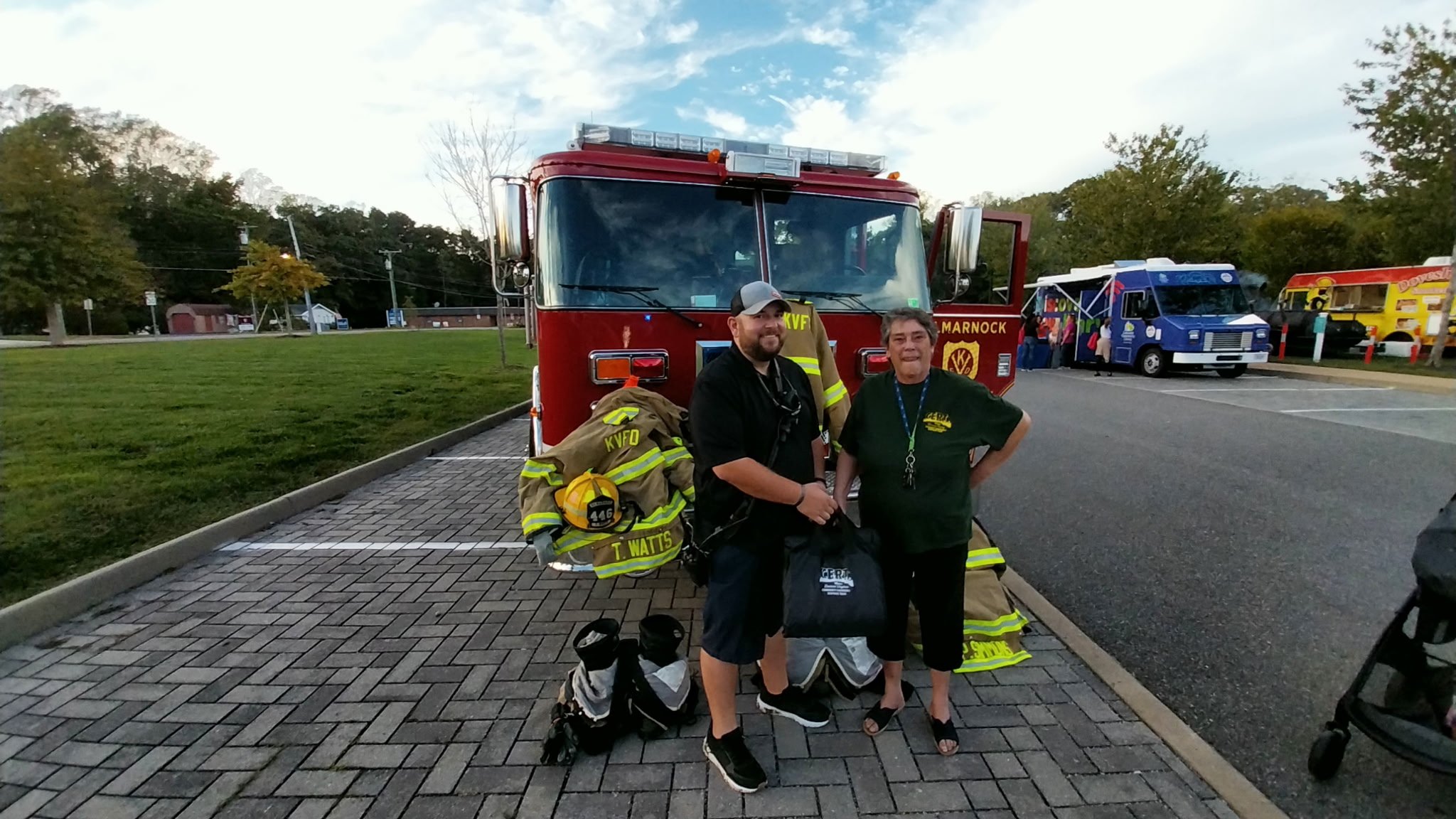 Two men standing in front of a fire truck and firefighters' gear, with food trucks and a grassy area in the background.