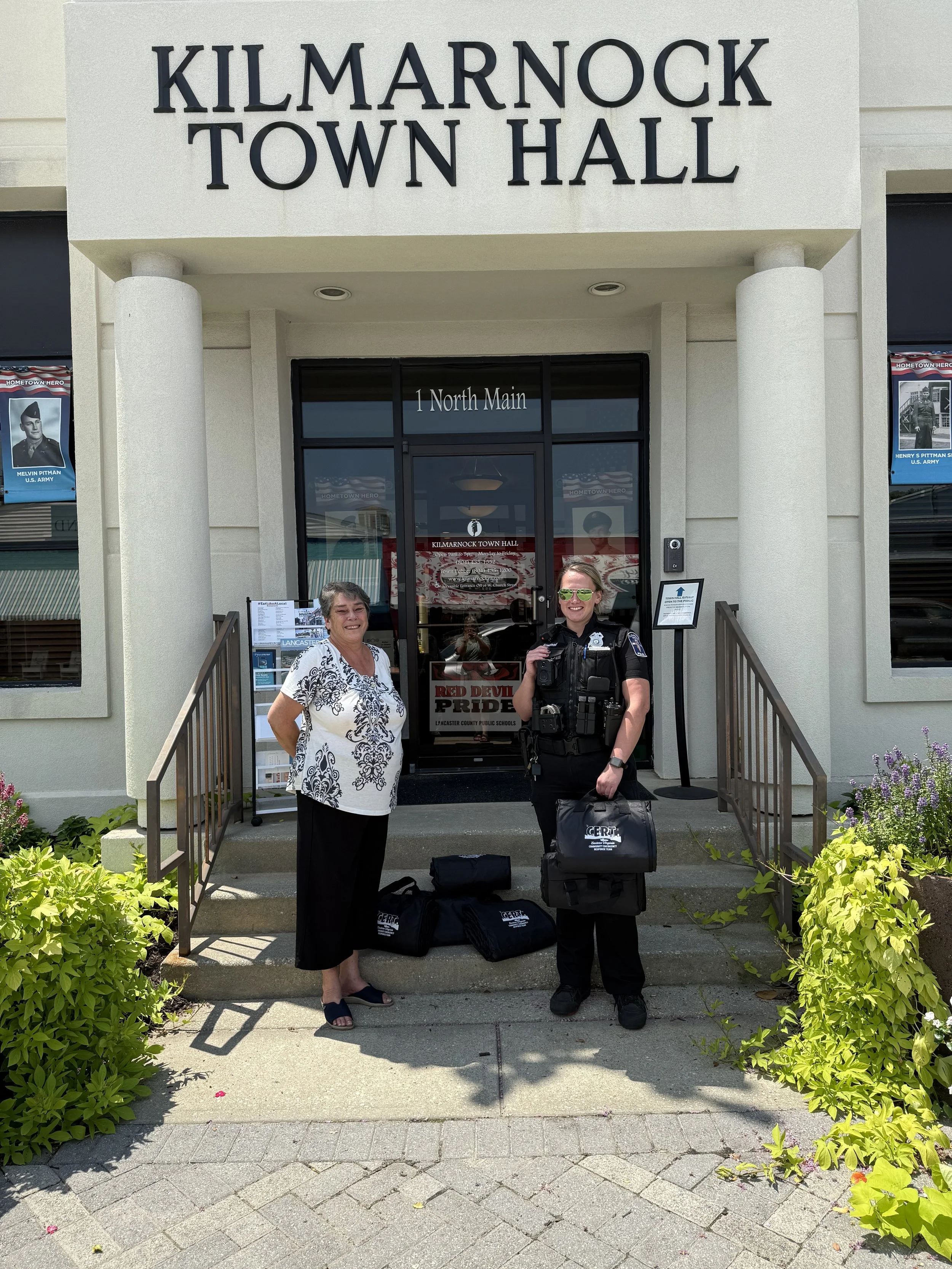 A woman and a police officer standing on the steps of Kilmarnock Town Hall, with plants on either side, bags on the ground, and posters visible in the windows.