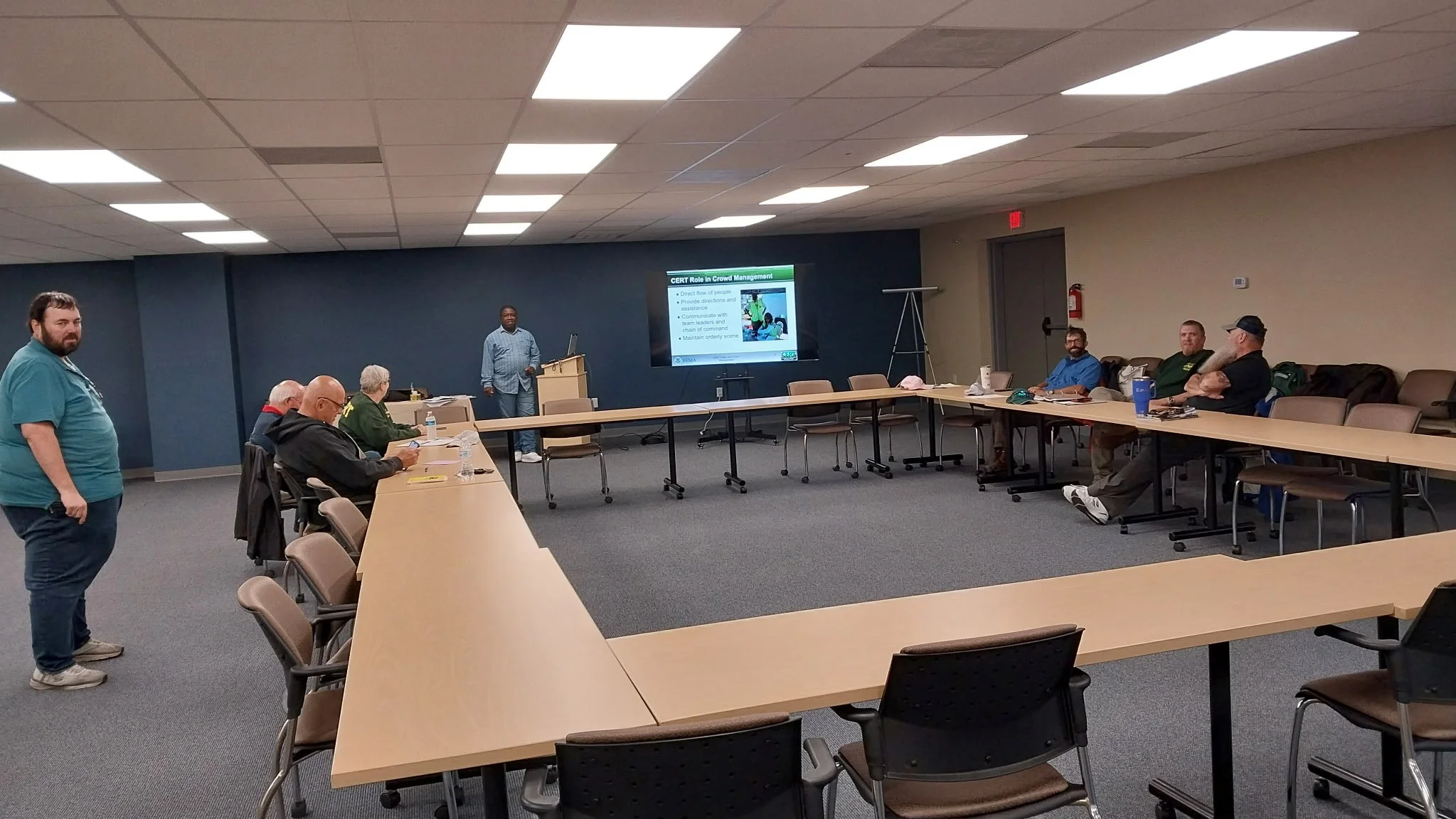 A conference room with a large U-shaped table surrounded by chairs, some of which are occupied. A man is standing at a podium giving a presentation on crowd management and CERT, with a projected slide behind him. Several attendees are seated around the table, some taking notes or looking at the presentation. The room has a drop ceiling with fluorescent lights, and a fire extinguisher is mounted on the wall to the right.