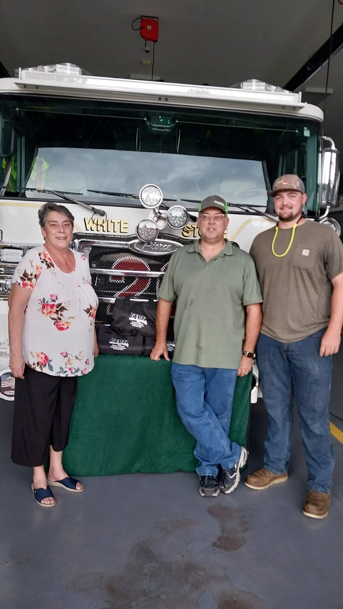 Three people standing in front of a fire truck inside a garage. The woman on the left is wearing a white floral top and black pants. The man in the middle is wearing a green shirt and blue jeans. The man on the right is wearing a brown shirt and jeans. The fire truck has the words "WHITE" and "ST" visible and is equipped with multiple lights and tools.